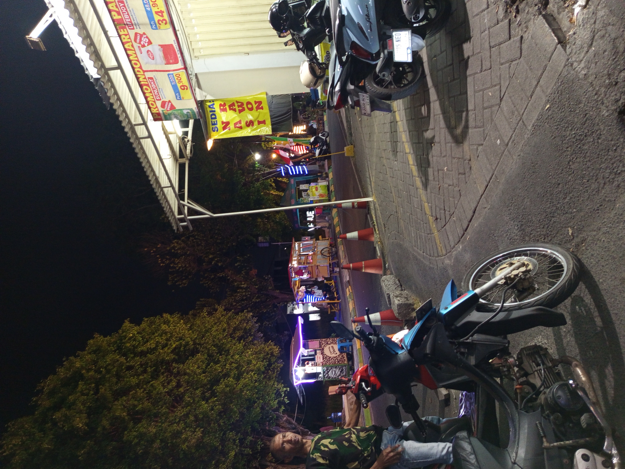 The image depicts a street scene at night in Gemurung, Indonesia. A man in a camouflage t-shirt and jeans stands near a parked motorcycle. In the background, several food stalls are illuminated with colorful lights, suggesting a vibrant night market atmosphere. One stall has a prominent yellow banner that reads "SEDIA RAWON NASI," indicating it sells rawon rice. Another stall displays promotional signs for Indomaret, a convenience store chain, with prices visible. Several other motorcycles are parked along the street, and traffic cones are placed on the side of the road. The ground is paved with bricks, and trees line the area. The overall scene conveys a lively commercial and social gathering space after dark.