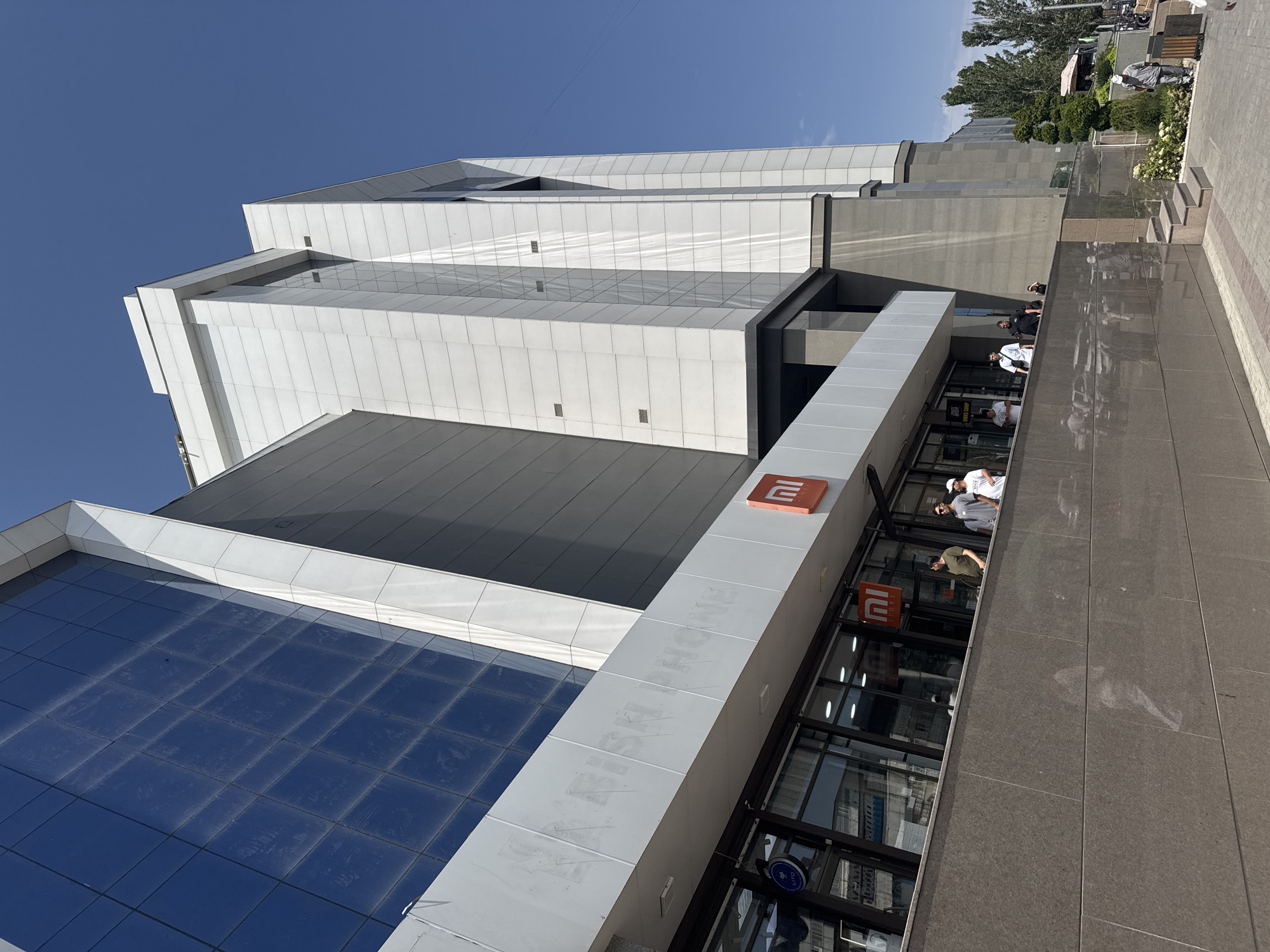 The image shows a modern building with a glass facade and white paneling, possibly a shopping mall or office complex. The building is under a clear blue sky. There are several people walking outside the building, some of whom appear to be customers or employees. A sign for "MI STORE" is visible, indicating a retail establishment selling Xiaomi products. The building is located in Bishkek City, Kyrgyzstan. The architecture is contemporary, with sharp angles and a mix of materials. The overall impression is that of a bustling urban environment on a sunny day.