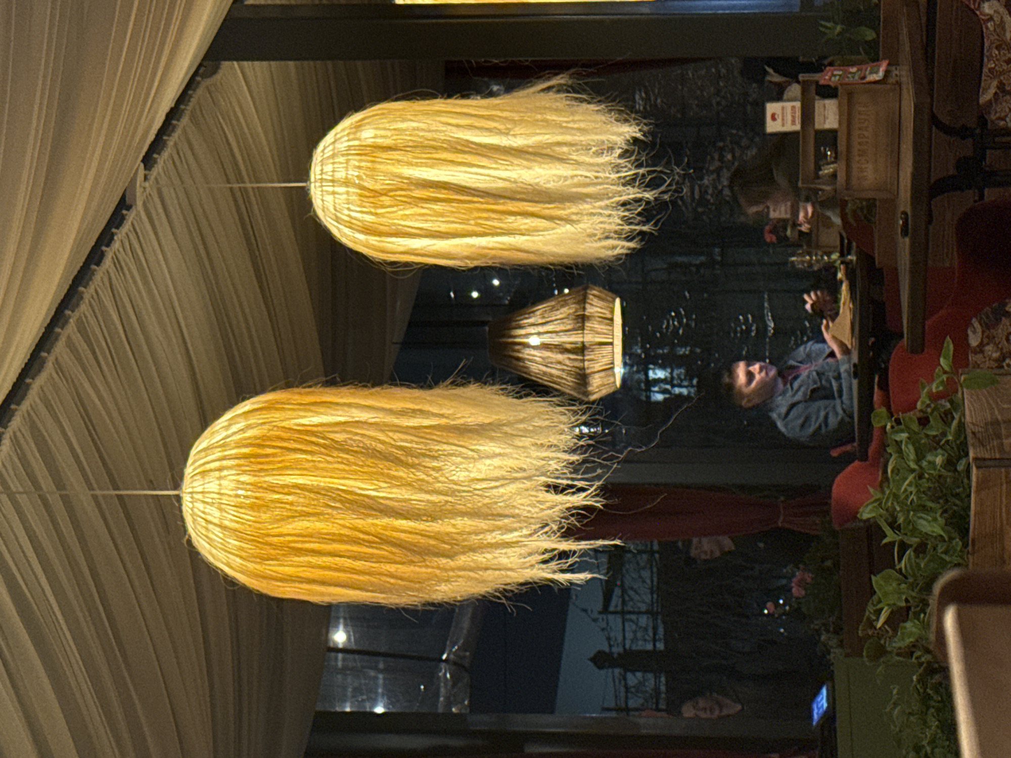 The image shows an interior scene in Perm, Russia. Two large, elongated, straw-colored pendant lamps hang from a beige draped ceiling. A smaller, similar-style lamp is visible further back. The background consists of a window reflecting a street scene. Inside, the reflection shows people seated at tables. A man in a blue jacket is visible. There are also decorative elements like wooden boxes and green plants. The furniture includes red chairs and wooden tables.