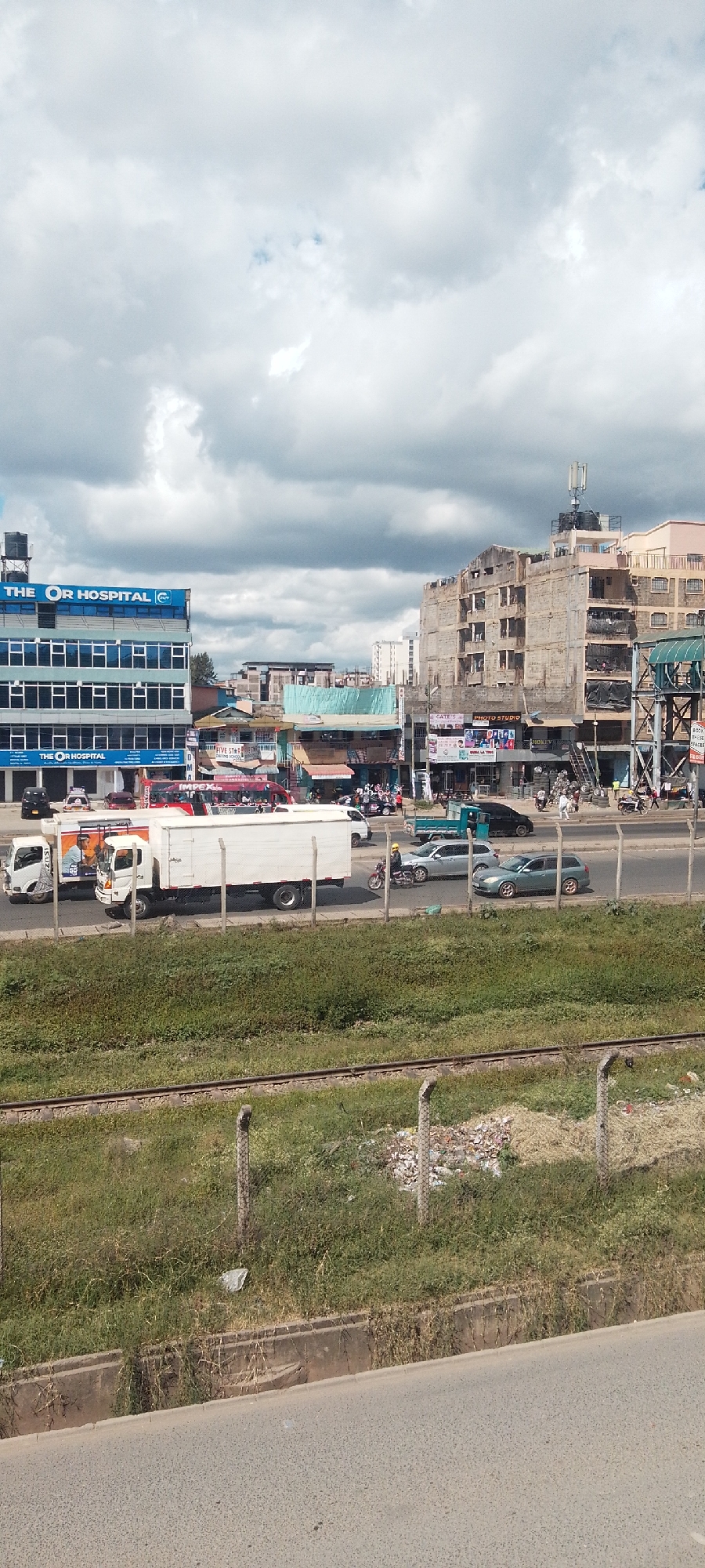 The image captures a busy street scene in Nairobi, Kenya. The foreground features a concrete barrier, green vegetation, and a section of railway tracks enclosed by wire fencing. Beyond this, a multi-lane road is filled with various vehicles including trucks, cars, and motorcycles, suggesting heavy traffic.
On the left side of the road, a modern building labeled "THE OR HOSPITAL" stands out, characterized by its blue and white color scheme. Further down the street, a collection of older, more densely packed buildings are visible, with commercial signage indicating various businesses, including what looks to be a photo studio. Above these buildings, telecommunications equipment, water tanks, and antennas are seen. The sky is overcast with thick, gray clouds. Pedestrians and other people can be seen walking.