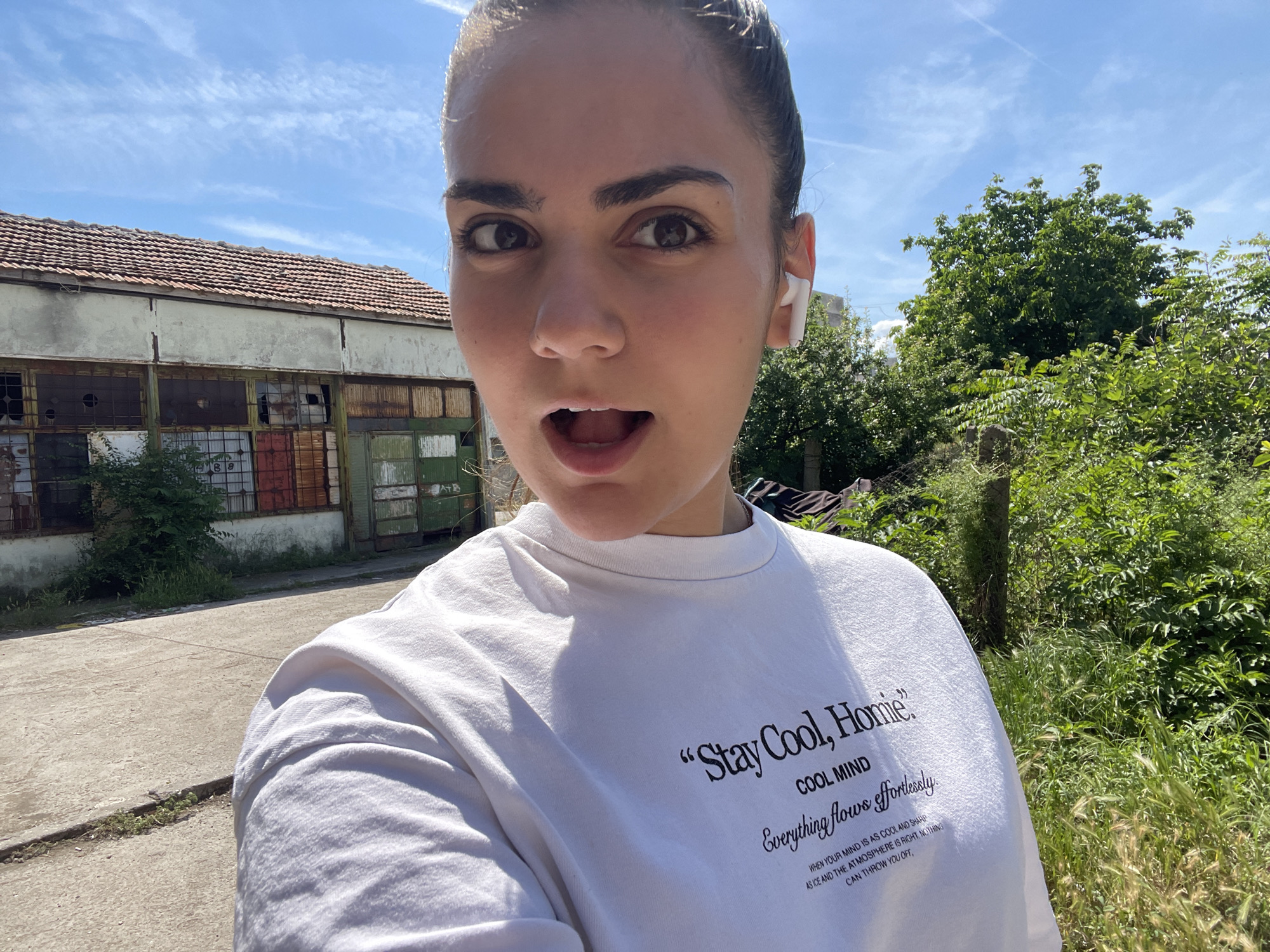 A woman with her hair pulled back, wearing a white t-shirt with text that reads "Stay Cool, Homie" and "COOL MIND, Everything flows effortlessly. WHEN YOUR MIND IS AS COOL AND SHARP AS ICE AND THE ATMOSPHERE IS RIGHT, NOTHING CAN THROW YOU OFF.", takes a selfie outdoors on a sunny day. She is wearing white earbuds. Behind her, on the left, is an old, weathered building with a tiled roof and broken windows. To the right of the building and behind the woman, there is lush green foliage. The ground in front of the building is paved concrete with some cracks. The location is the city of Debelt, Bulgaria.