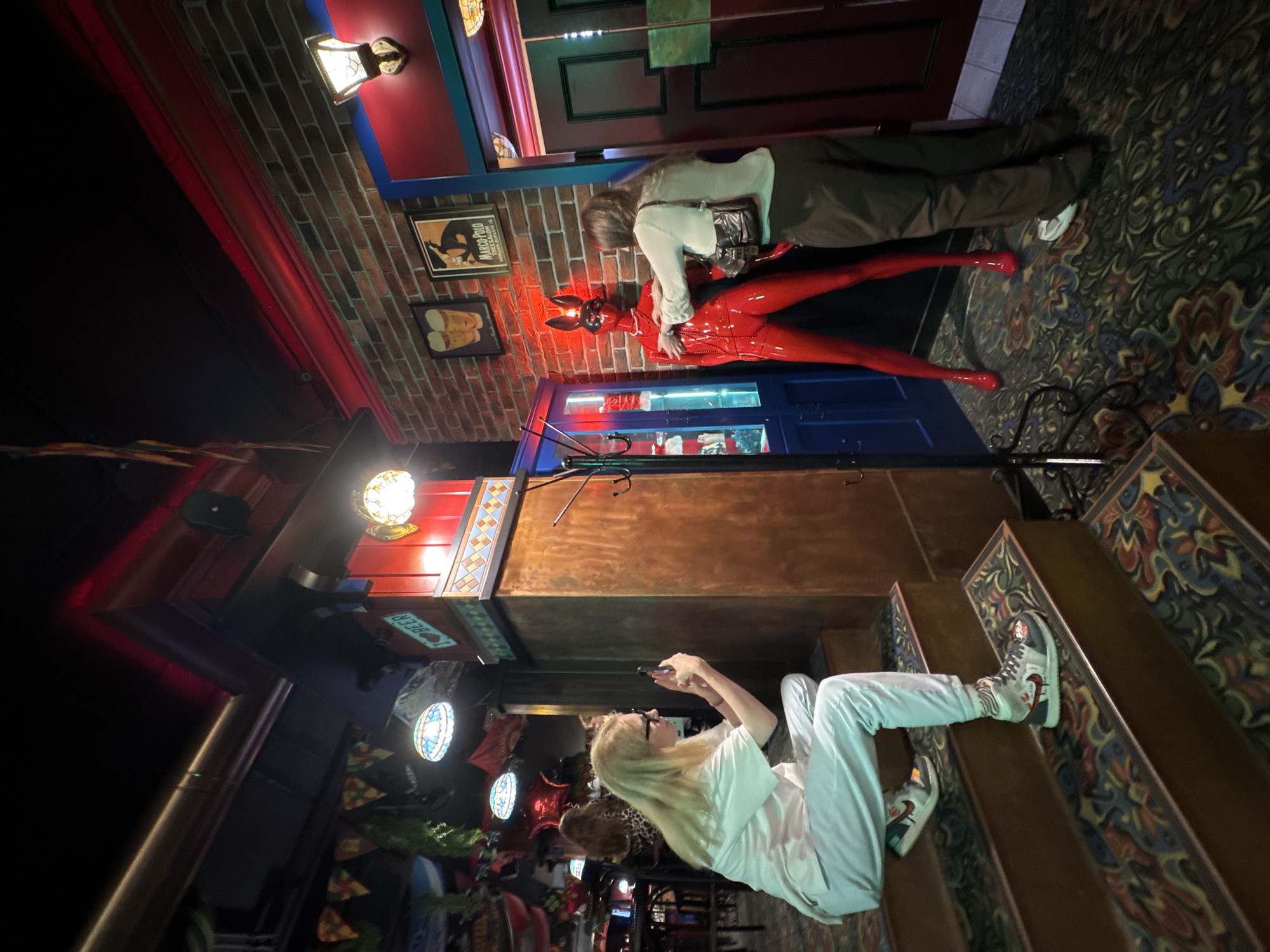 The image shows an indoor scene, possibly a bar or club, with an eclectic decor. In the foreground, a person with blonde hair is sitting on wooden stairs, taking a picture with their phone. They are wearing a white t-shirt and light-colored pants, and athletic shoes. Further into the scene, another person is standing and appears to be interacting with a mannequin dressed in a red, shiny full-body suit and a black mask with rabbit ears. The mannequin is positioned next to a blue-trimmed doorway. The environment features brick walls, decorative lighting fixtures, and patterned carpeting. There are also various decorative items and posters visible in the background. The image does not contain any information that would allow for the identification of the city of Izhevsk, Russia, or any other specific location.