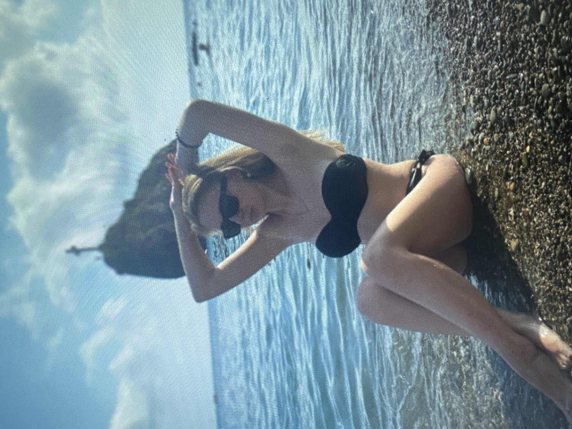 In this medium shot, a young woman is seated on a pebble beach, with her legs submerged in the shallow, clear blue water. She is wearing a black bikini and sunglasses, and her arms are raised behind her head, with her hands resting on her blonde hair. The background features a rocky outcrop with a statue at its summit, and a bright, cloudy sky. The water is calm and reflects the light, creating ripples on its surface. The overall impression is one of a relaxed and serene beach scene.