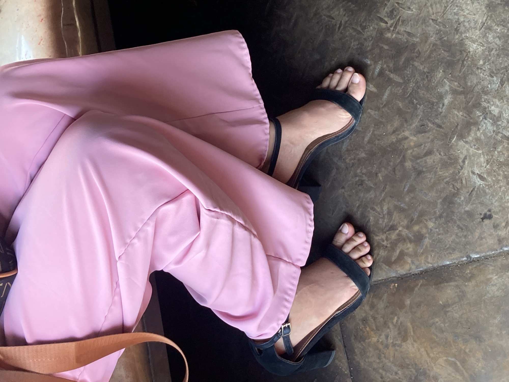 The image shows a close-up view of a person's legs and feet, clad in pink wide-leg pants and black open-toed heeled sandals. The sandals have a single strap across the toes and an ankle strap. The person is seated, with their legs crossed. The background appears to be a textured, possibly metallic or concrete surface, suggesting an urban or industrial setting. The lighting is natural and slightly diffused, indicating it might be daytime. No specific landmarks or indicators within the image can be used to pinpoint the location to Davao City, Philippines.