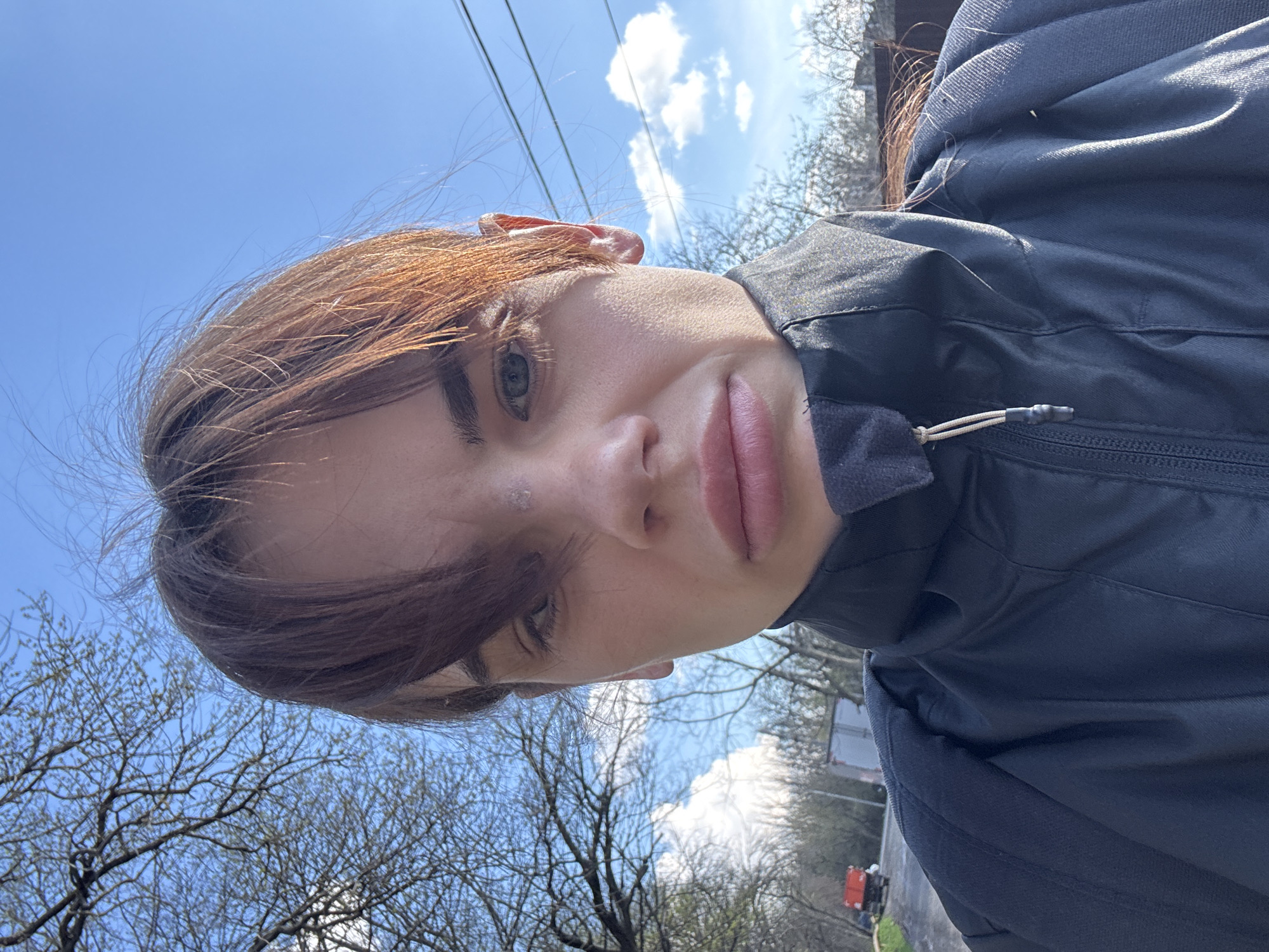 A low-angle outdoor selfie of a young fair-skinned person with reddish-brown hair and green eyes, wearing a dark jacket, possibly in Rostov-on-Don, Russia (due to the request for location information). The background includes bare trees, a blue sky with some clouds, and overhead electrical wires. In the distance, there is some urban development visible, including what appears to be a small road.