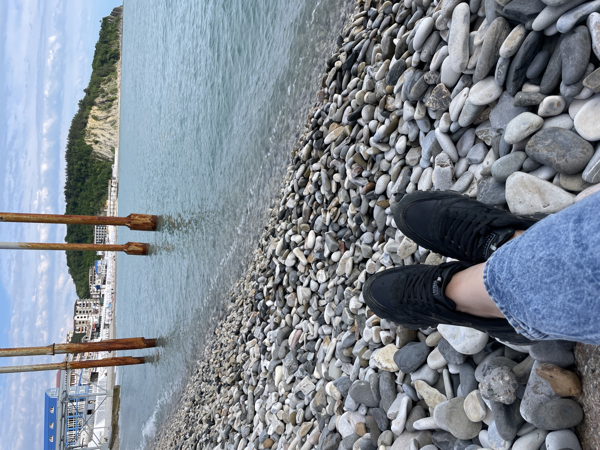 The image depicts a view from a pebble beach in Olginka, Russia. A pair of black sneakers on the beach and light blue jeans are visible. The beach is made up of various smooth, rounded pebbles. Calm seawater is washing the edge of the beach. In the background, there are rusty metal poles and a section of buildings. A cliff covered in greenery is further in the distance. The sky is partly cloudy.