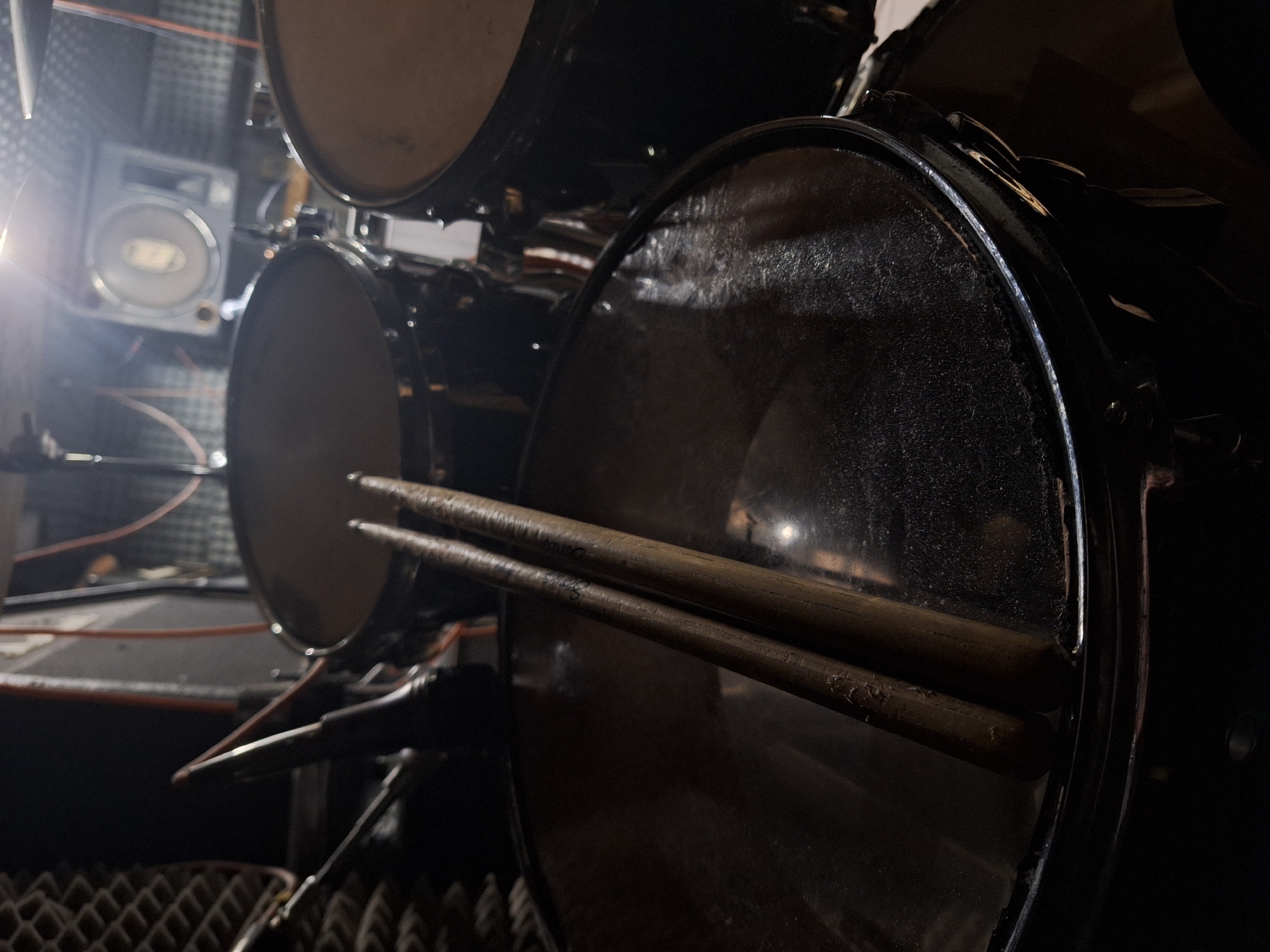 A close-up, low-light photograph captures parts of a drum kit set up in what appears to be a music studio or practice room in Panagyurishte, Bulgaria. In the immediate foreground, two worn wooden drumsticks rest horizontally across the dusty, reflective head of a large drum. The drum head shows signs of use and has a slightly grimy texture.
Behind the prominent drum, another drum head is partially visible in the mid-ground, also appearing dark and somewhat worn. To the far left, a bright, intense light source creates a strong flare and illuminates a speaker or amplifier mounted on a wall. The speaker has a circular cone and some visible controls, though any text or branding on it is obscured by glare and blur. The walls in the background and foreground exhibit acoustic foam panels, characterized by their pyramidal or egg-crate texture, designed to absorb sound. Various wires and cables are subtly present in the background, hinting at the setup of musical equipment.
The overall scene is dimly lit, with deep shadows contrasting sharply with the bright light source on the left. The condition of the equipment suggests it is actively used. No people are visible, and no specific activity beyond the implied readiness for music is taking place. There is no clearly legible text on any of the visible objects. The atmosphere evokes a sense of quiet anticipation within a dedicated musical space.