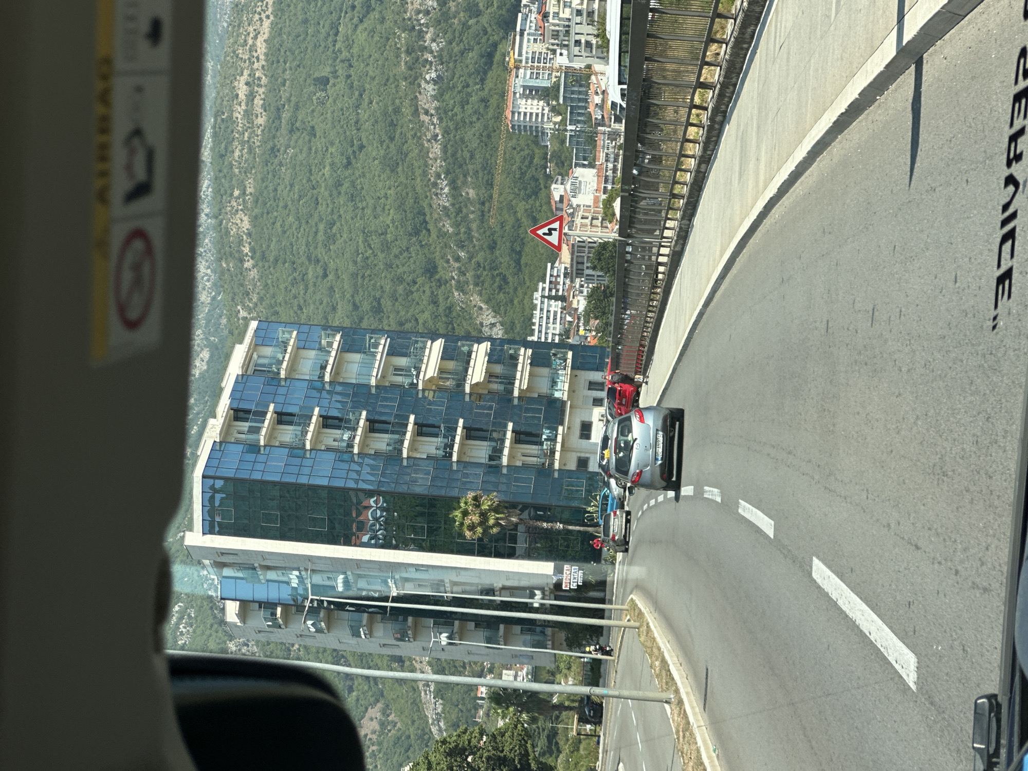 The image, taken from a vehicle's perspective in Boreti, Montenegro, shows a street scene with a modern glass-fronted building on the left, a road with several cars, and a lush green hillside in the background. The building is tall and features multiple balconies with white railings and awnings. A palm tree stands near its base. The road curves to the right, with white lane markings. Cars visible include a silver hatchback with a license plate that reads "90-AN974", a red car, and a grey car. A motorcyclist is also present on the left side of the road. Further down the road, a triangular traffic sign with a winding road symbol is visible. Beyond the road, a metal fence lines a pedestrian walkway, and a cluster of buildings with red-tiled roofs and a construction crane are nestled against the wooded hillside. The sky is bright and sunny. The image also shows a portion of the vehicle's interior, including the top edge of the windshield and what appears to be an airbag warning label.