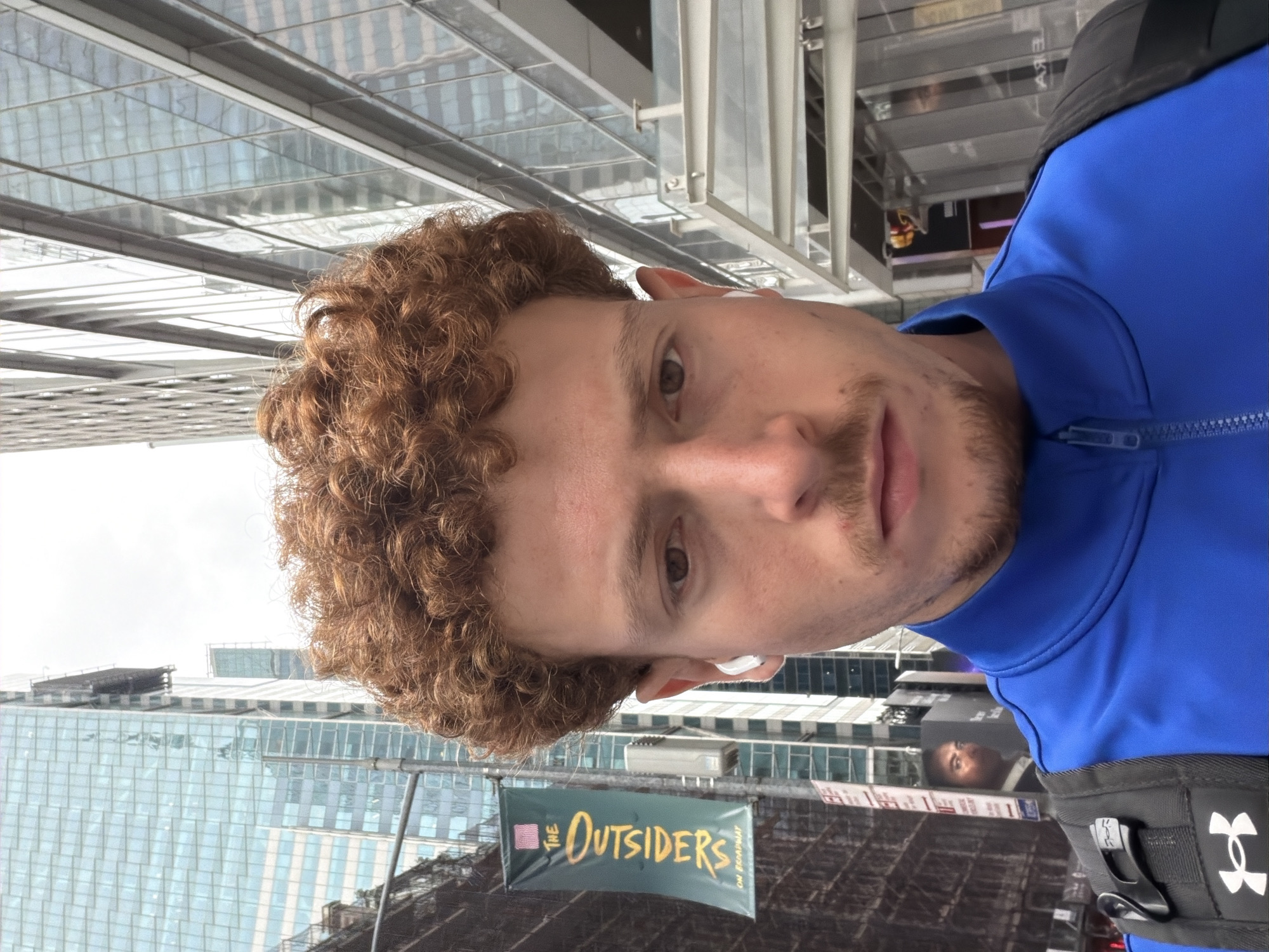 The photograph shows a man with curly auburn hair and light skin. He is wearing a blue jacket and a black Under Armour bag. He is standing in front of a banner that reads "The Outsiders on Broadway." Behind him are skyscrapers. This appears to be in the Times Square area of New York City.