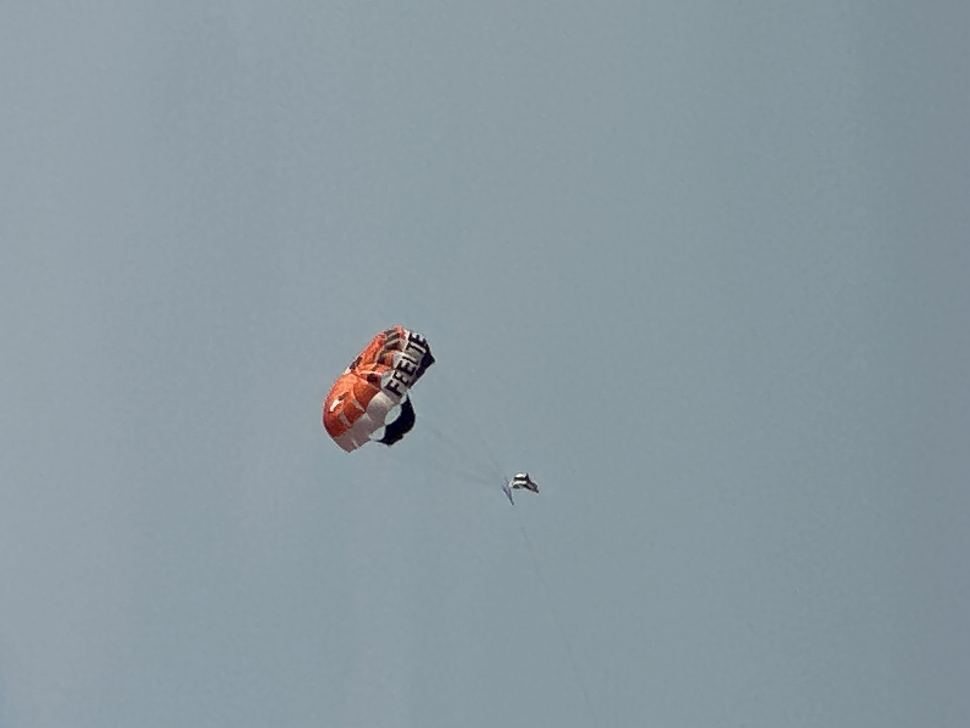 An orange and white kite with the word "FEEL" printed in black letters is depicted against a pale blue sky. A small, dark object, possibly a person on a paraglider or hang glider, is visible in the lower left quadrant of the image, tethered to the kite by thin lines. The kite appears to be a stunt kite or parafoil kite, and it is positioned to the right of the small object. The background is a clear, unobscured sky, suggesting an outdoor activity in an open space. No urban elements or specific landmarks of Boreti, Montenegro are visible in this image.