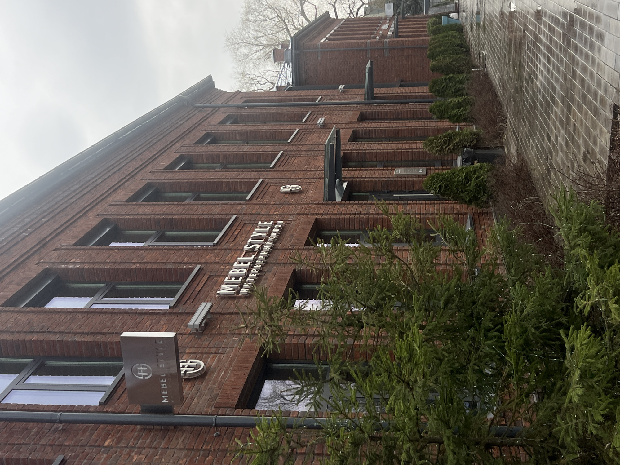 A tall, multi-story building constructed of reddish-brown brick dominates the frame, viewed from a low angle on an overcast day. The building features numerous rectangular windows with dark frames, set into recessed sections of the brick facade, giving the brickwork a textured, stepped appearance between levels.
Prominently displayed on the building is a sign reading "MEBEL STYLE" in metallic, raised Latin script. Below this, "ИНТЕРЬЕРНЫЙ САЛОН" (meaning "INTERIOR SALON" in Russian) is written in Cyrillic. A stylized logo, resembling intertwined letters, is visible on this sign and on a separate circular plaque mounted on the building.
In the foreground, dense green coniferous trees or bushes with prominent needles partially obscure the lower and middle sections of the building. To the right, a paved or tiled path runs parallel to a low, light-colored brick wall, in front of which a row of neatly trimmed evergreen bushes is planted. The ground, especially the paved area, appears damp, suggesting recent rain or wet conditions. Bare tree branches are silhouetted against the cloudy, light gray sky in the upper background. The scene depicts an urban or commercial building, likely located in Kazan, Russia, under diffused daylight conditions.