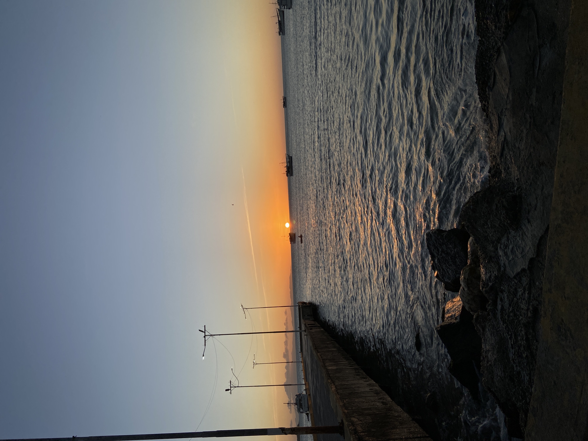 A vertical photograph captures a sunrise over the ocean in Juan Griego, Venezuela. The sun is positioned on the horizon, casting a golden glow across the water. Several boats are visible in the distance on the water. A concrete pier extends into the water, lined with lampposts. The pier is made of concrete and is visible at the bottom half of the photograph. Rocks are visible on the right side of the photograph. The sky transitions from a light blue to a vibrant orange.
