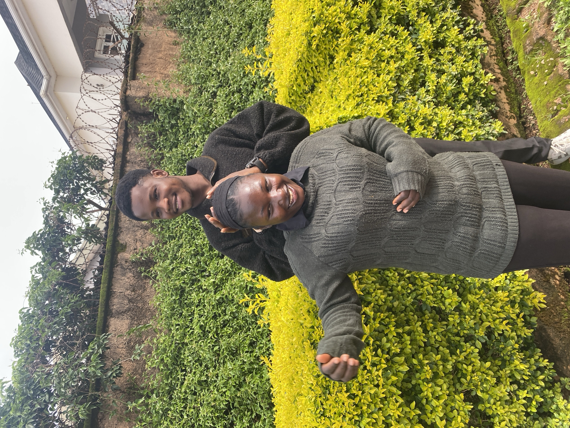 The image shows two young adults, a man and a woman, posing for a photo outdoors. They are in a seated position, with the man behind the woman, his hands resting on her head. Both are smiling broadly, appearing happy and affectionate.
The man is wearing a dark, textured sweater or jacket and has short, dark hair. The woman is wearing a dark, knitted sweater and a black head covering. Her eyes are closed, and she has a wide, joyful smile.
They are situated in a garden setting with lush green bushes and some vibrant yellow foliage behind them. A wall is visible to the left, with barbed wire on top, suggesting a secured area. Above the wall, part of a building with a grey roof is visible, along with some trees. The overall impression is that of a casual, outdoor portrait taken on a bright day, possibly during daylight hours, given the lighting. The scene evokes a sense of warmth and connection between the two individuals. The location context of Jos, Nigeria is not directly indicated by visual cues in the image itself. There is no visible text in the image.