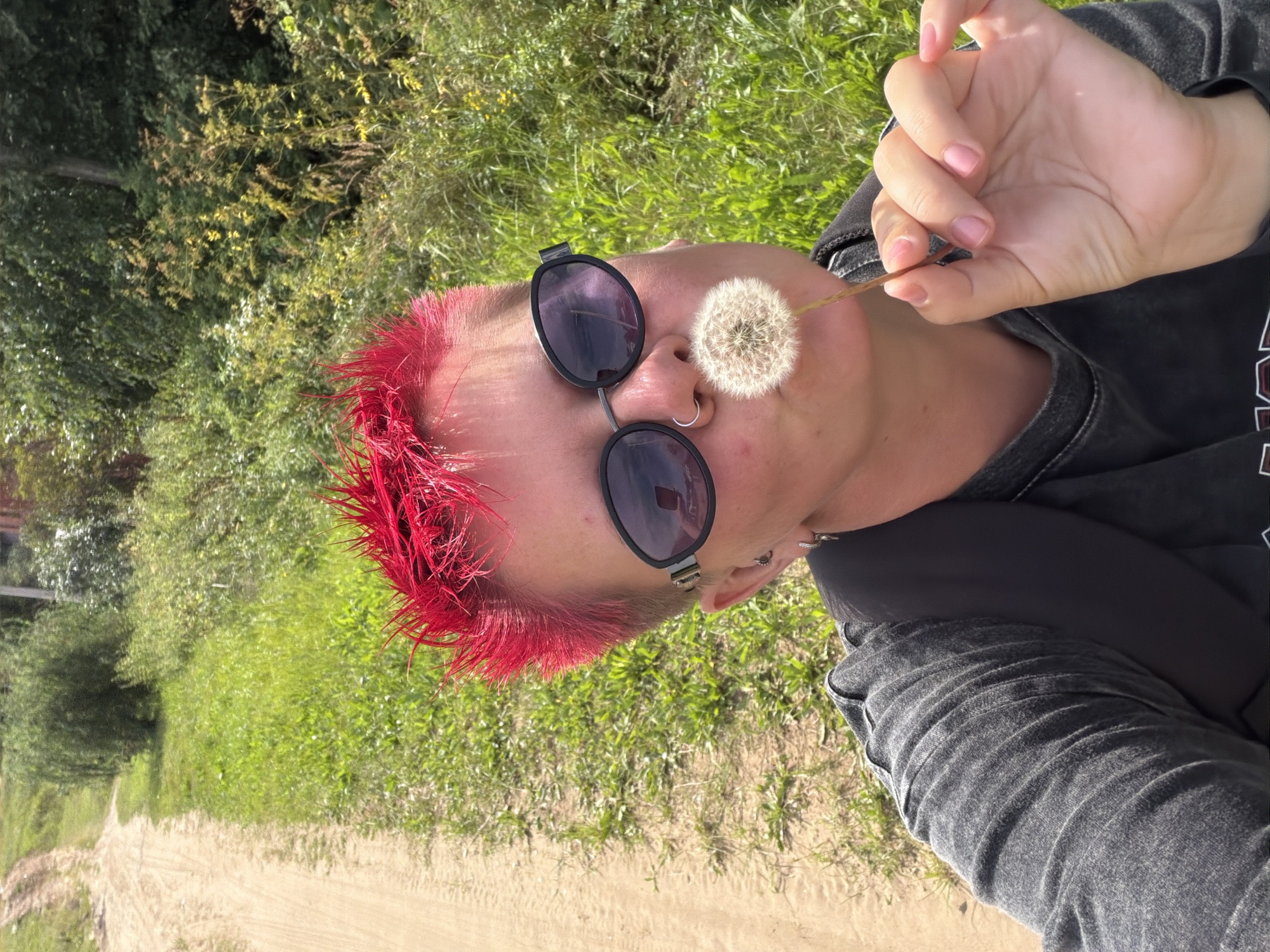 In the city of Chaikovsky, Russia, a person with bright red, spiky hair and sunglasses is holding a dandelion puffball up to their mouth, as if to blow it. They are wearing a dark grey t-shirt and a black backpack. The background consists of a dirt road on the left and lush green foliage and trees on the right, suggesting an outdoor, rural or park-like setting. The lighting indicates it is daytime.