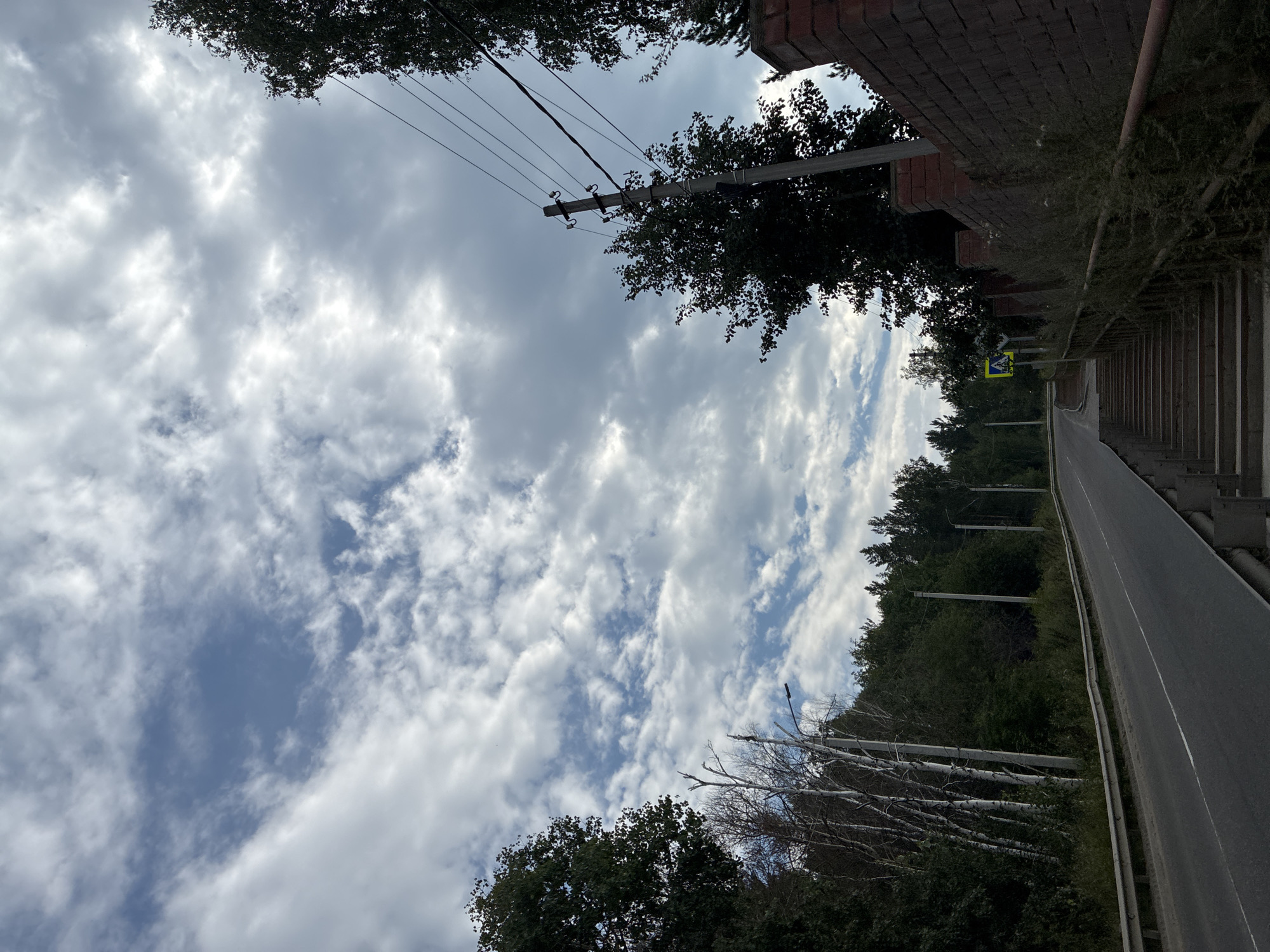 The image depicts a cloudy sky over a road and a pedestrian walkway with stairs. The road curves to the left and appears to be in a rural or semi-rural setting, bordered by trees. There is a metal guardrail along the road. To the right of the road, a brick wall runs parallel to it, and beyond that are trees and a utility pole with wires. A set of stairs leads down from the road level, with a handrail and overgrown vegetation. A pedestrian crossing sign is visible in the distance on the left side of the road. The sky is filled with dramatic clouds, with patches of blue sky visible through them. The overall impression is of an overcast day in a natural environment near a road. The location is specified as Markova, Russia.
