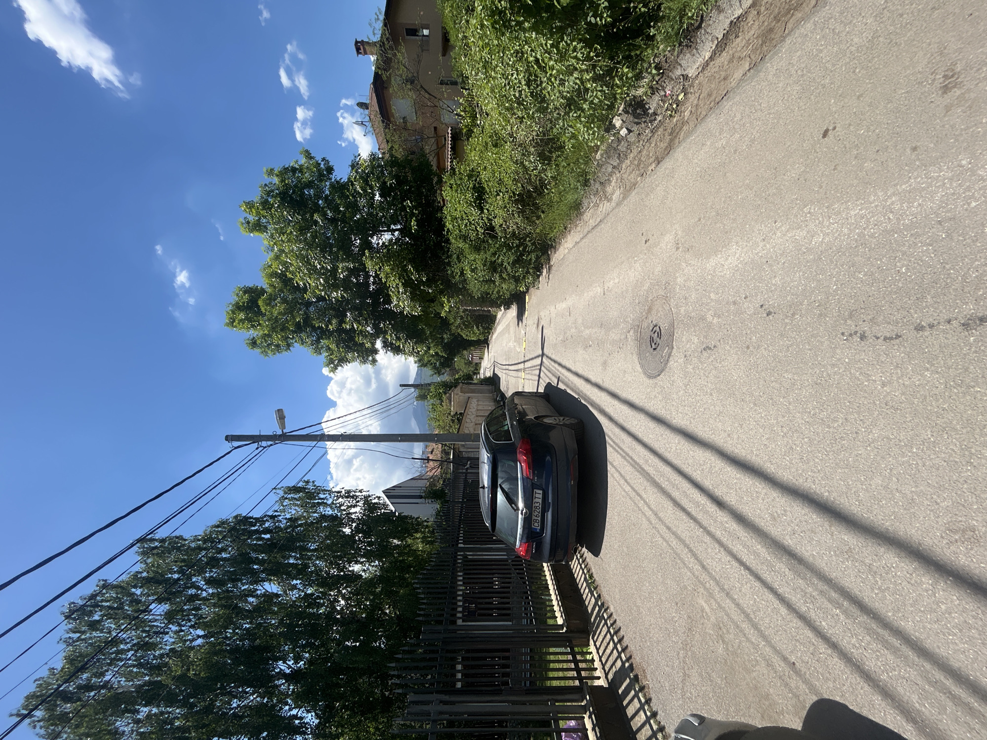 The image shows a street in Sofia, Bulgaria, with a blue Opel Insignia station wagon parked on the right side of the road. The car's license plate number is CB 6283 TT. The road is paved and has a manhole cover on the surface. Shadows of power lines stretch across the road. A black metal fence runs along the left side of the road, with trees and bushes behind it. A building with a red-tiled roof is visible in the background on the right. The sky is blue with scattered clouds. A power pole with a streetlight is also visible on the left side of the road. The location appears to be a residential area.