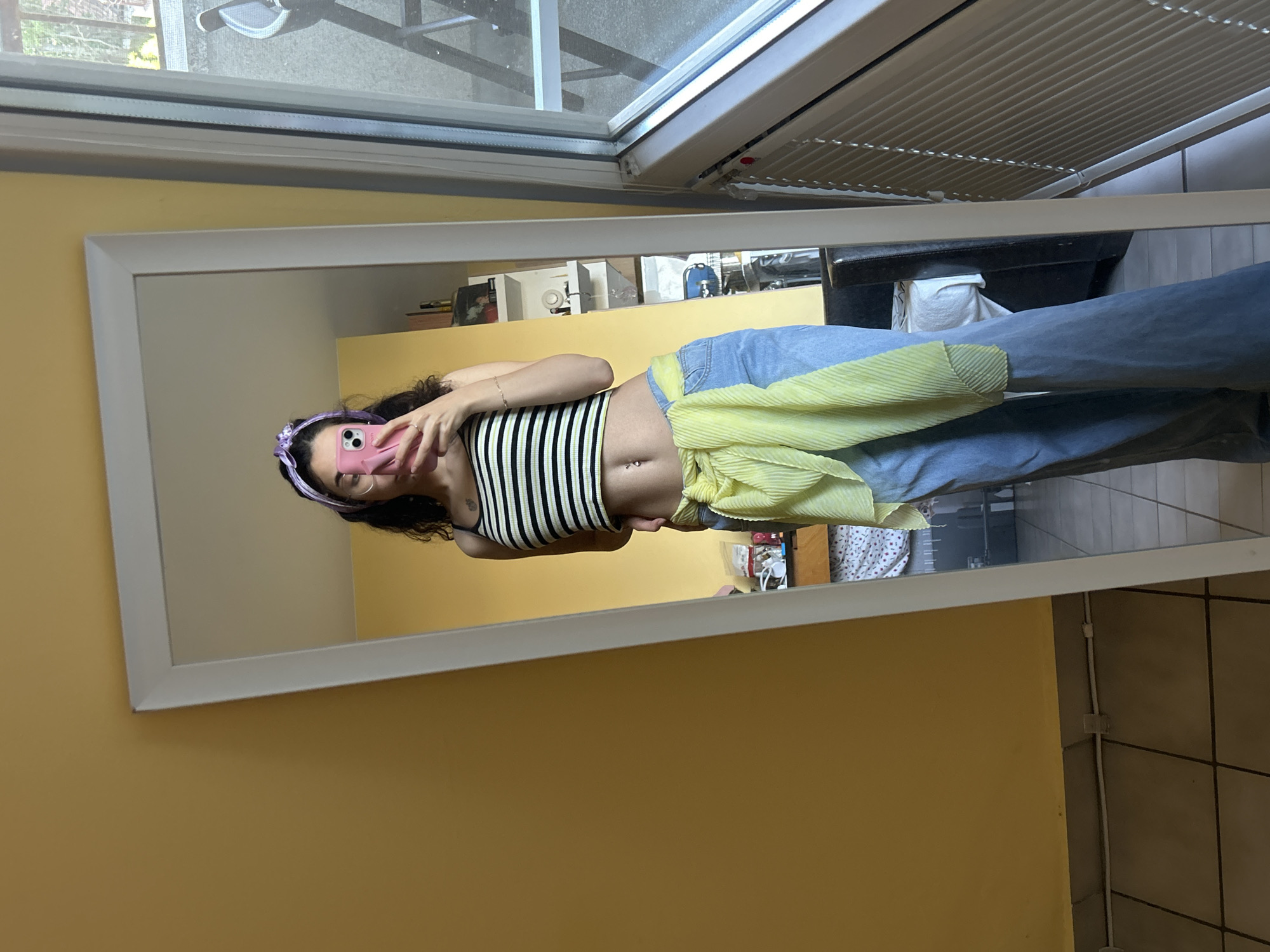 A person is taking a selfie in a mirror. The mirror is leaning against a yellow wall. The person is wearing a striped top, jeans, and a yellow shirt tied around their waist. There is a window in the background. There is no information to determine the exact location in Sofia, Bulgaria.