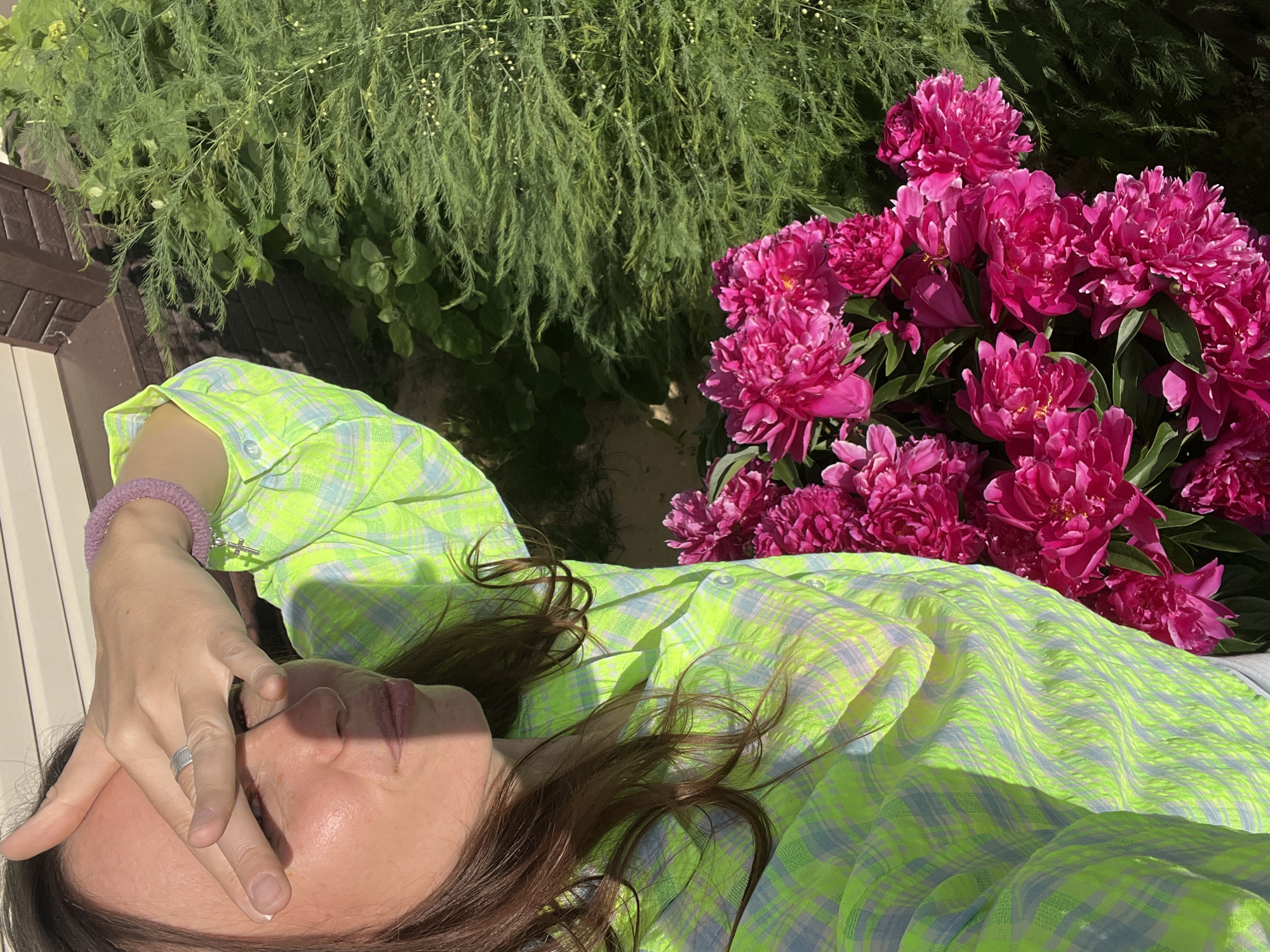A young woman is taking a selfie outdoors, in front of some foliage and a house. She is wearing a bright green and blue checkered shirt and has her hand over her eyes to block out the sun. A ring is visible on her finger. To the right of the woman, a large bouquet of bright pink peonies is visible. The background shows green foliage, a portion of a house, and a textured surface that could be part of the house's exterior. The image appears to have been taken during the day, with strong sunlight casting shadows. This scene is located in Pigozino, Russia.