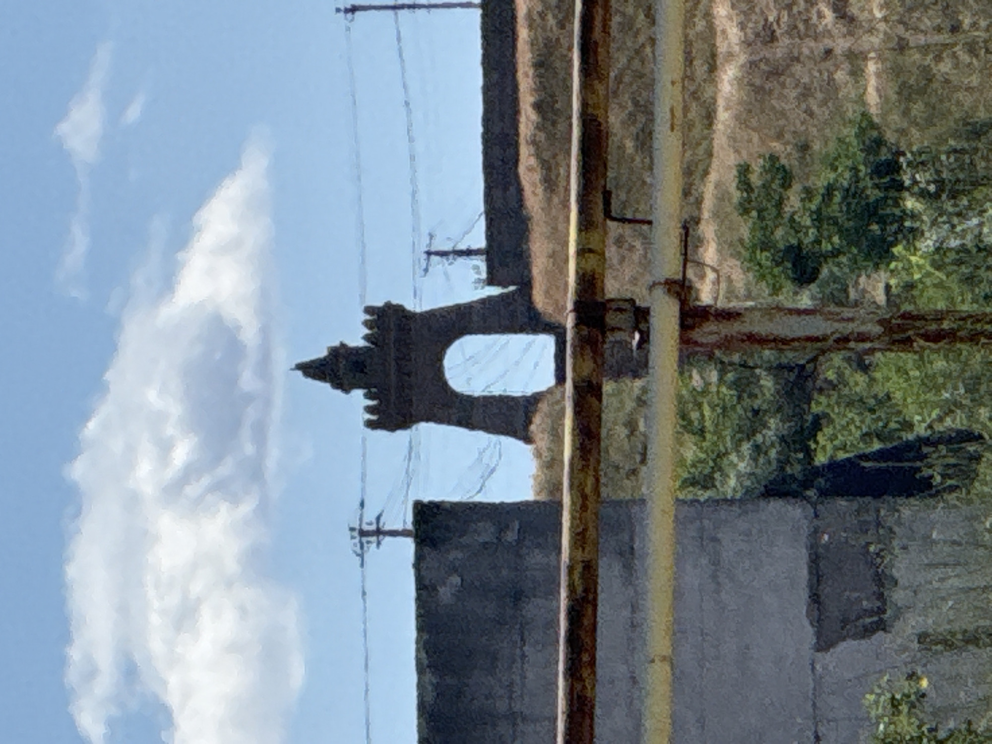 The image captures a view through a rusty, yellow-painted metal railing towards a stone archway with a crenellated top, resembling a small castle turret. The archway is centered in the background, framed by two dark, rough-textured walls. In the distance, a line of power poles and wires stretches across the frame against a bright blue sky with a fluffy white cloud. The area surrounding the archway appears to be a dry, grassy slope. In the foreground, the railing is prominent, with a dark gray concrete structure to the left and green foliage to the right, suggesting an outdoor, possibly semi-urban or rural setting. The image is taken from a slightly low angle, looking upwards towards the archway and the sky.