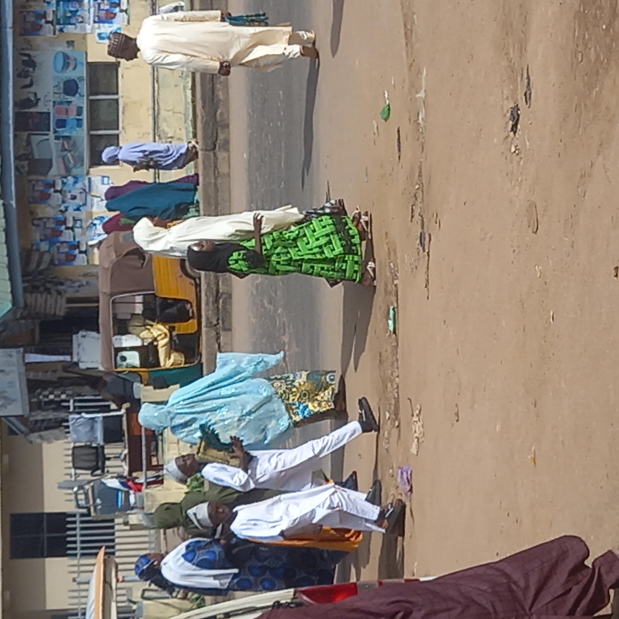 A street scene in Dadi Riba, Nigeria, shows a line of people walking or standing near a road. Several people are wearing traditional Islamic clothing, including hijabs and robes. A yellow auto-rickshaw is parked near the sidewalk. Debris litters the ground. Buildings are visible in the background.