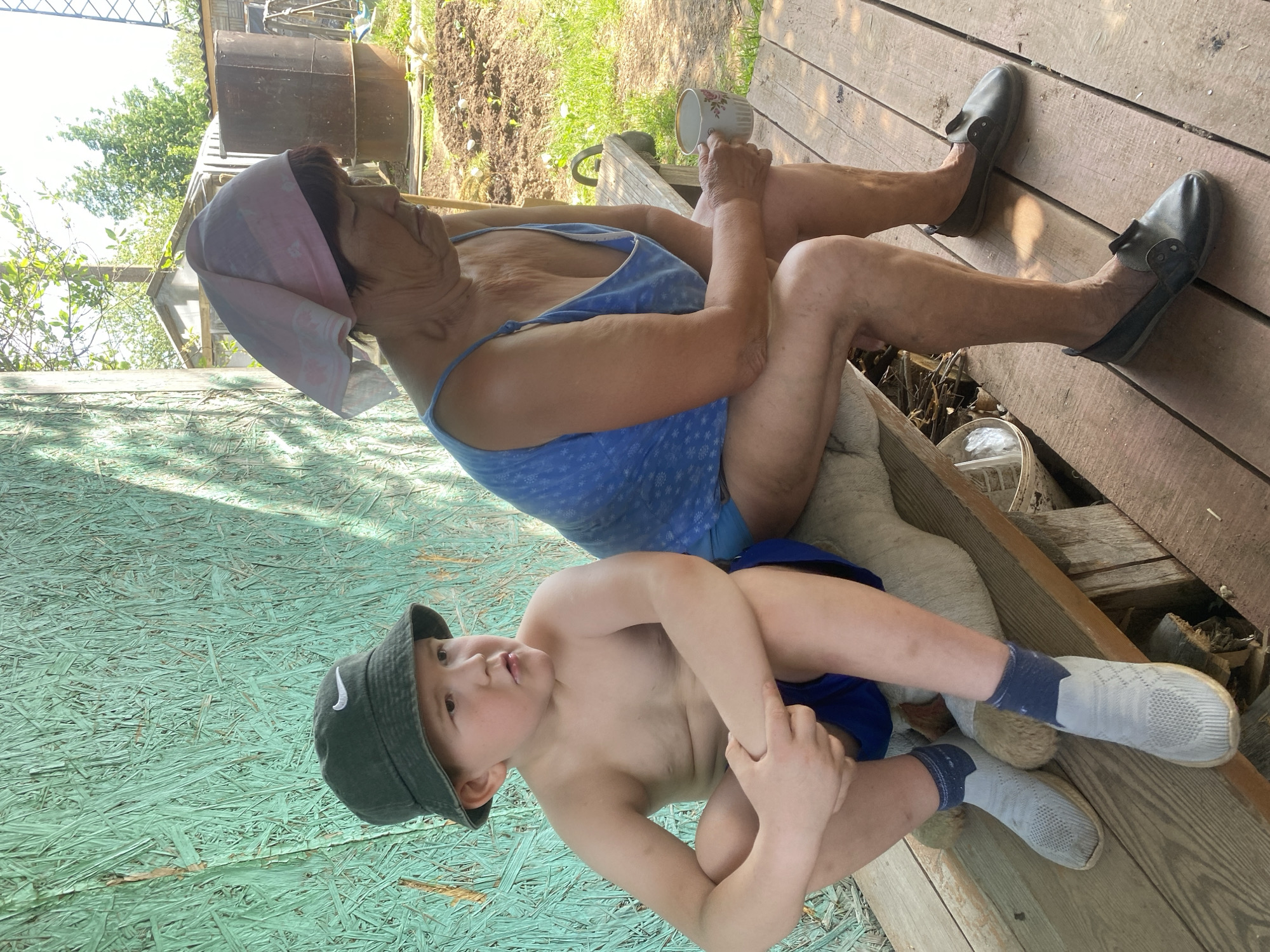 The image depicts an elderly woman and a young boy seated outdoors on a wooden structure. The woman wears a blue dress with a white floral pattern and a pink headscarf, and she is holding a white cup with a floral design. The boy is shirtless, wearing blue shorts and a green Nike bucket hat, and he has on grey socks and shoes. The scene is set in what appears to be a garden or backyard, with some greenery visible in the background. The structure they are sitting on includes light green painted plywood and weathered wood planks. The location is Прикамский, Russia.
