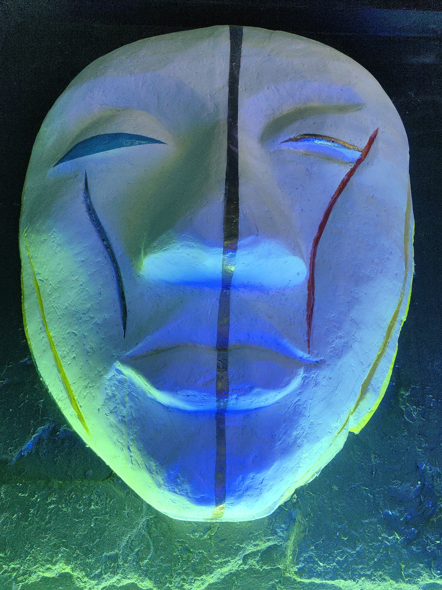 A close-up, head-on view of a white, stylized mask is presented, illuminated by vibrant green and blue lights. The mask, likely made of plaster or clay, has a rough, textured surface. It depicts a serene, almost mournful expression with closed eyes and full lips. A prominent vertical black line bisects the mask down the center, extending from the forehead to the chin. A thin red line runs down the right side of the mask from the eye area, resembling a tear or stylized marking. The mask is framed by a dark background, with the lower portion of the image showing a textured, rocky surface bathed in an intense green light. The lighting creates a dramatic and somewhat otherworldly atmosphere, with blue light illuminating the face and green light highlighting the surroundings. The mask appears to be an object of art or cultural significance, possibly displayed in a darkened space. No specific activity or event is depicted. The setting could be an exhibition, a performance space, or an outdoor art installation. The location context provided is Baan Mae Nam, Thailand, which might suggest the mask has a cultural or traditional relevance to the region.