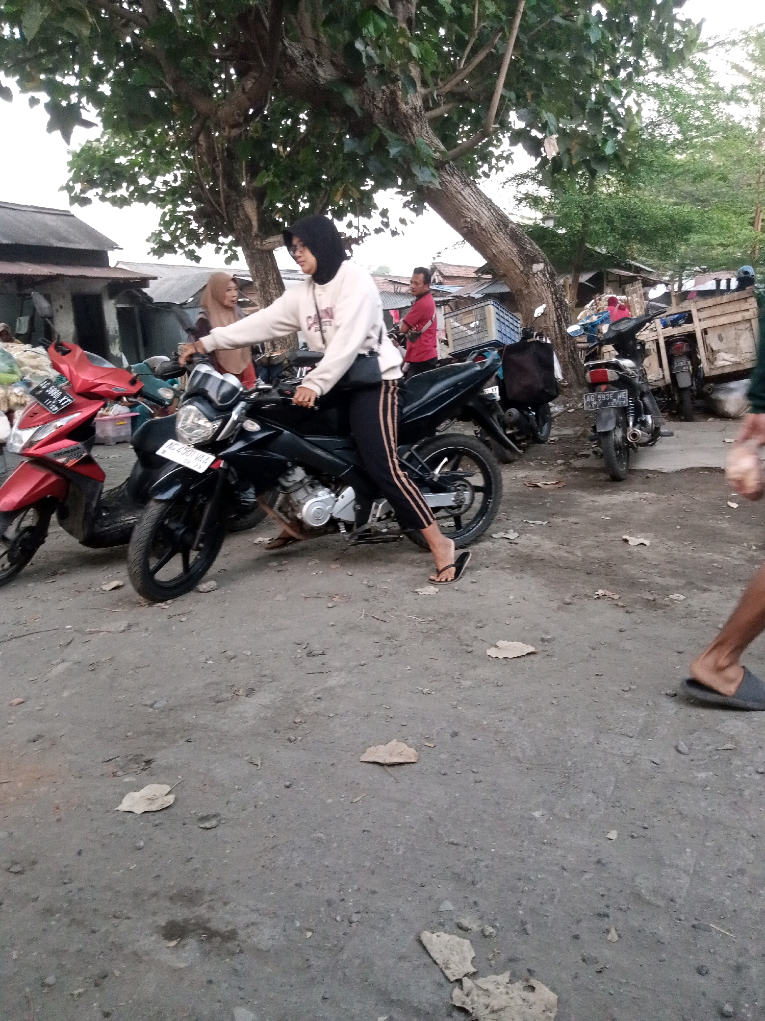 A medium shot shows a woman on a motorcycle in an outdoor market in Watu Dandang, Indonesia. The woman, wearing a hijab and a cream-colored sweatshirt, is seated on a black motorcycle with a license plate reading "AG 2501 VAA". She is holding the handlebars with her right hand and appears to be looking forward. To the left of her motorcycle is a red scooter. In the background, several buildings with corrugated metal roofs are visible, along with trees and other motorcycles. The ground is a dirt and gravel surface, with scattered leaves. The overall scene suggests a busy, informal marketplace.