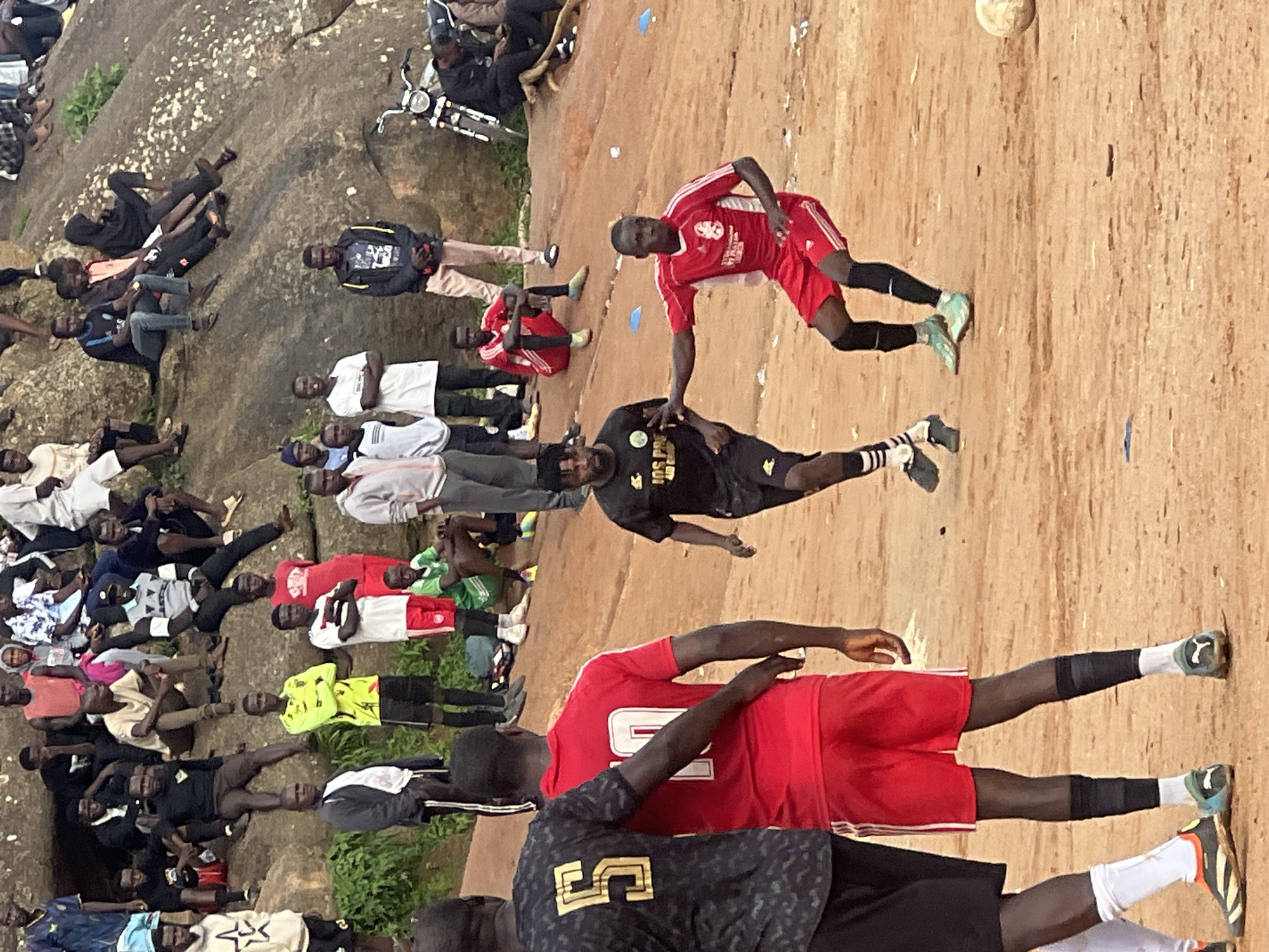 The media file depicts a group of young men playing football in a dusty, outdoor area in Jos, Nigeria. The scene is characterized by a rough, sandy ground, with a large rock formation visible in the background, suggesting a natural or semi-developed area. Several players are actively engaged in the game, with one player in a red jersey appearing to be kicking the ball, which is in mid-air. Other players are scattered around the field, some watching, others in various stages of anticipation or movement.
The clothing worn by the individuals varies, with many in athletic attire, including jerseys, shorts, and football boots. Some are in casual wear. The lighting suggests it is daytime, likely afternoon, with no clear indication of specific weather conditions beyond the dusty environment. The overall mood seems to be one of casual sport and camaraderie.
Notable details include a motorcycle parked near the rock formation and some scattered blue and white objects on the ground, possibly markers or debris. There is no visible text on the clothing of the players that is legible enough to be deciphered. The image captures a moment of lively football activity in a community setting.