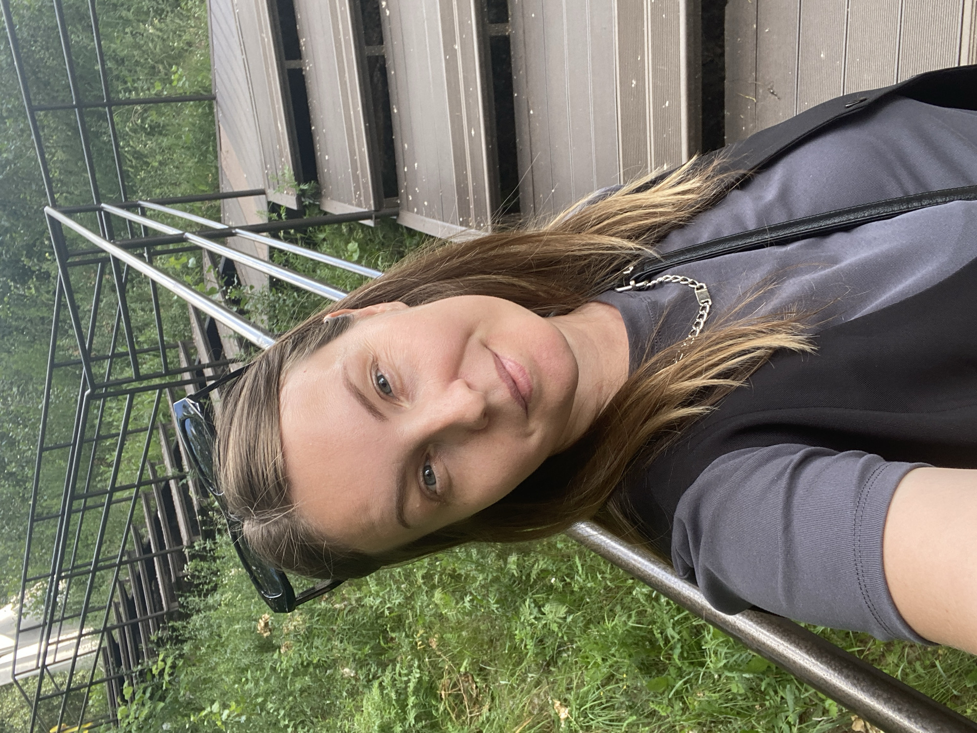 The media file is a portrait-oriented, eye-level selfie of a woman outdoors. She has long, light brown hair and fair skin. She is wearing a grey t-shirt with a black vest over it, and a silver chain necklace. Dark sunglasses are perched on top of her head. The woman is smiling gently at the camera.
The background features a set of wooden bleachers or outdoor seating, with metal railings. To the left, there is a metal fence and lush green grass and foliage, suggesting a park or natural setting. The lighting indicates it is daytime, likely with mild weather, as the woman is dressed casually. The overall mood is relaxed and pleasant. There is no visible text in the image. The context provided places the scene in Chaikovsky, Russia.