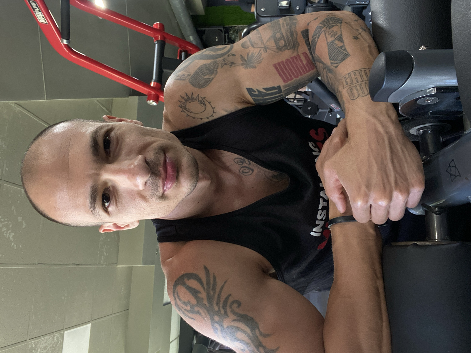 The image shows a man with tattoos on his arms and head, posing in a gym. He is wearing a black tank top and leaning on a piece of gym equipment. The gym appears to be well-equipped with various machines. The lighting suggests an indoor setting. The background shows the gym's interior, including other equipment and possibly other patrons in the distance. The man's expression is neutral, and he is looking directly at the camera. The overall impression is a portrait of a fitness enthusiast in a gym environment. This gym is located in Pleven, Bulgaria.