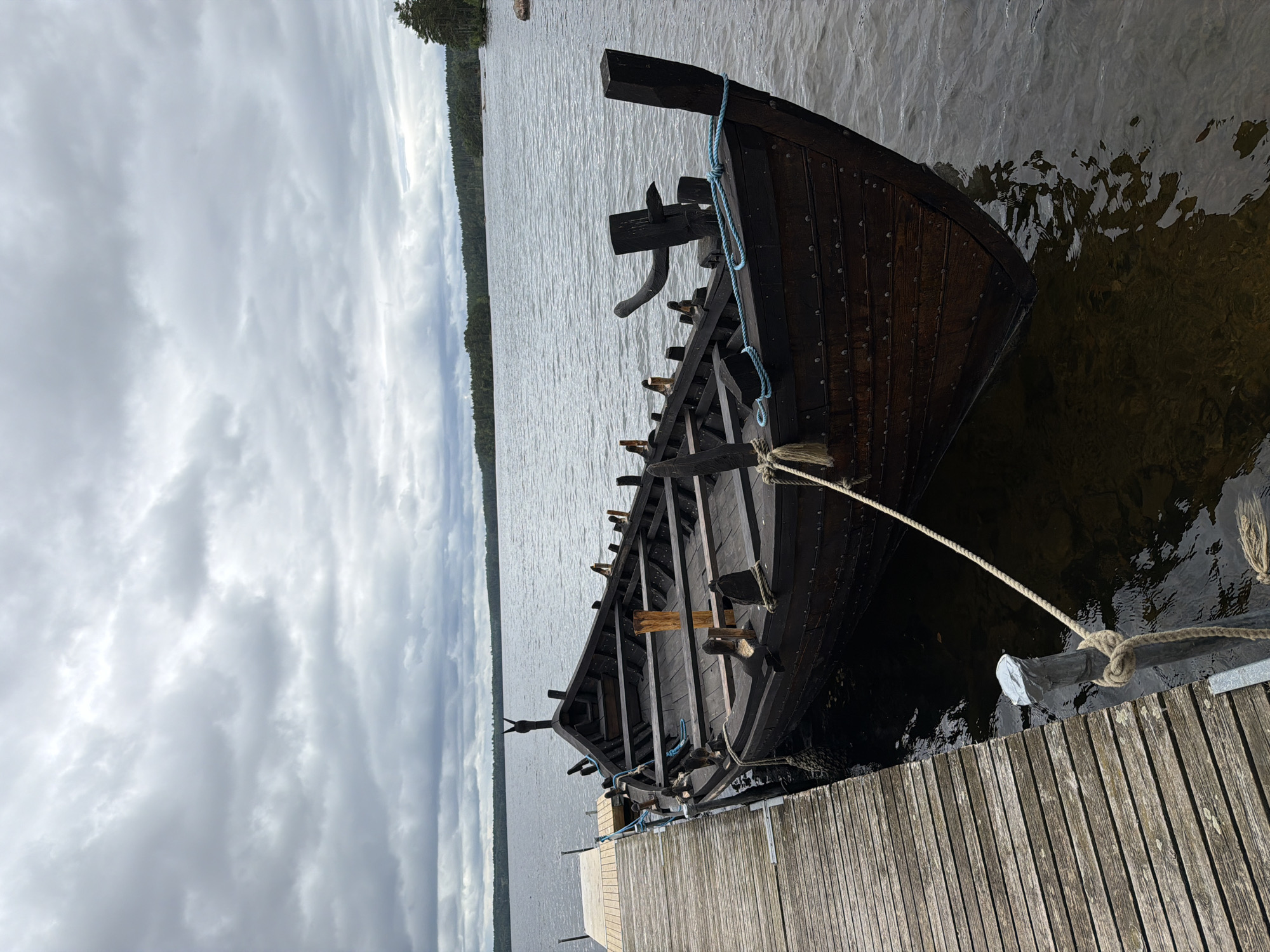 A dark wooden longboat is moored to a wooden pier on a calm lake under an overcast sky. The longboat has a pointed bow and stern and appears to be of historical design, possibly a replica. It is secured by ropes to posts on the pier. The water is dark and still, reflecting the cloudy sky. In the background, a tree-lined shore is visible across the lake. The scene suggests a quiet, natural setting in Aneby, Sweden.