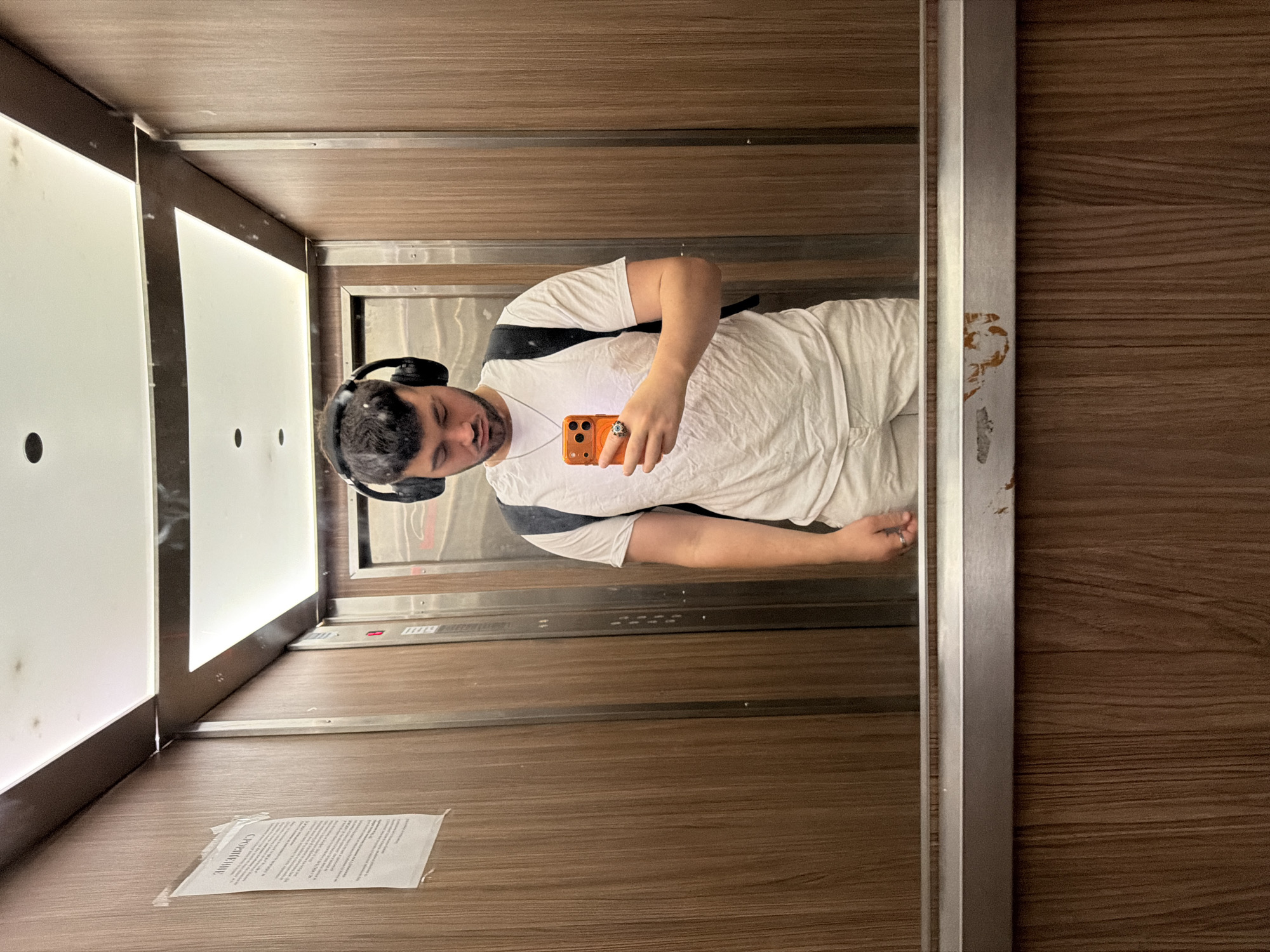 In the city of Sofia, Bulgaria, a man takes a mirror selfie inside an elevator. He is wearing a white t-shirt, white shorts, and black headphones. He is holding an orange smartphone in his right hand and a ring is visible on his left hand. The elevator has wood-paneled walls and a metal railing. A poster is affixed to the left wall of the elevator. The ceiling of the elevator is illuminated by recessed lighting fixtures. The floor of the elevator is not visible. The reflection in the mirror shows the man's face and upper body.