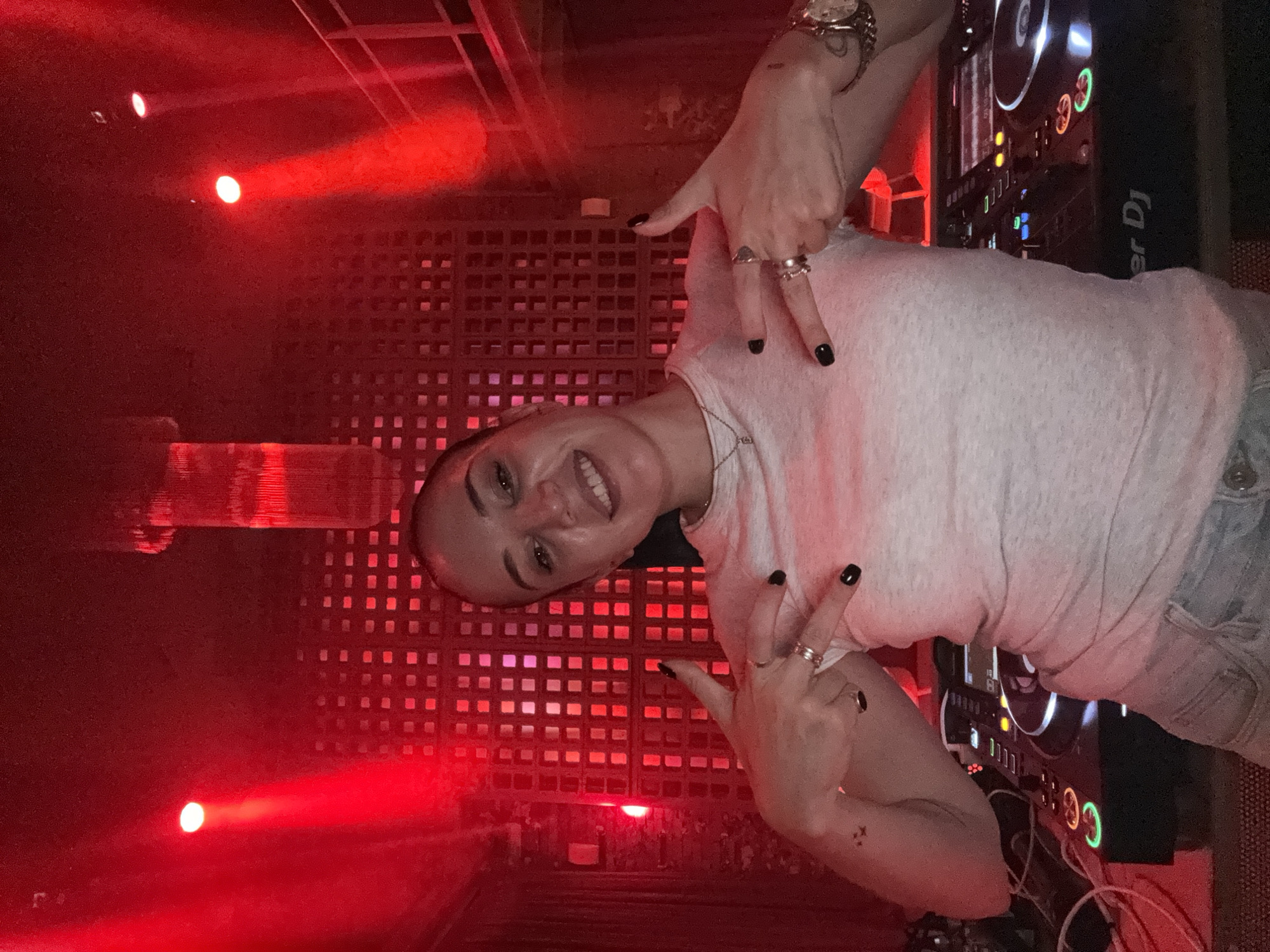 A woman is positioned in front of a DJ setup, making a hand gesture. She has dark hair pulled back from her face, a light-colored shirt, and a wristwatch on her wrist. Her fingernails are painted black. The DJ equipment to her right includes turntables and mixers. The environment is lit with a red hue, with red spotlights visible in the background. A wall with a patterned grid design is behind her. The location appears to be a nightclub or similar entertainment venue, potentially in Moscow, Russia based on the context, though specific markers are not visible to definitively confirm the city.