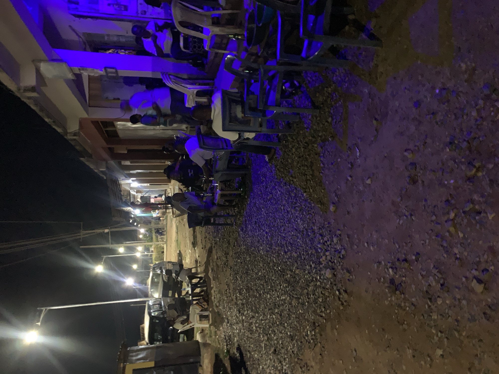 The image shows an outdoor scene at night in what appears to be Kabong, Nigeria. People are seated on chairs arranged along a building's exterior. Streetlights illuminate the area. The ground is a mix of dirt and gravel. Cars are parked nearby. A blue light source casts a distinct hue in some areas.