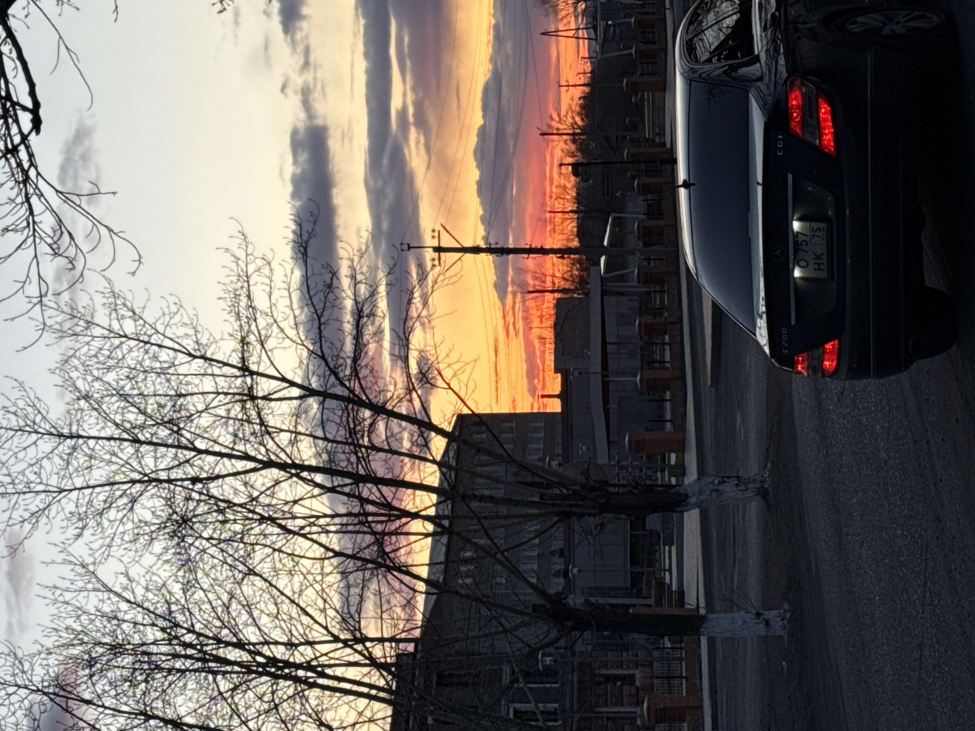The image depicts a black Mercedes-Benz C200 CGI parked on a street in Borzya, Russia. The license plate number is O 757 HK 75. The background features a sunset with orange and pink hues in the sky, visible through bare trees. Buildings and power lines are also visible in the background, suggesting an urban or industrial area. The foreground shows the street with a paved surface. The scene is captured from a slightly low angle, looking upwards towards the car and the sky.