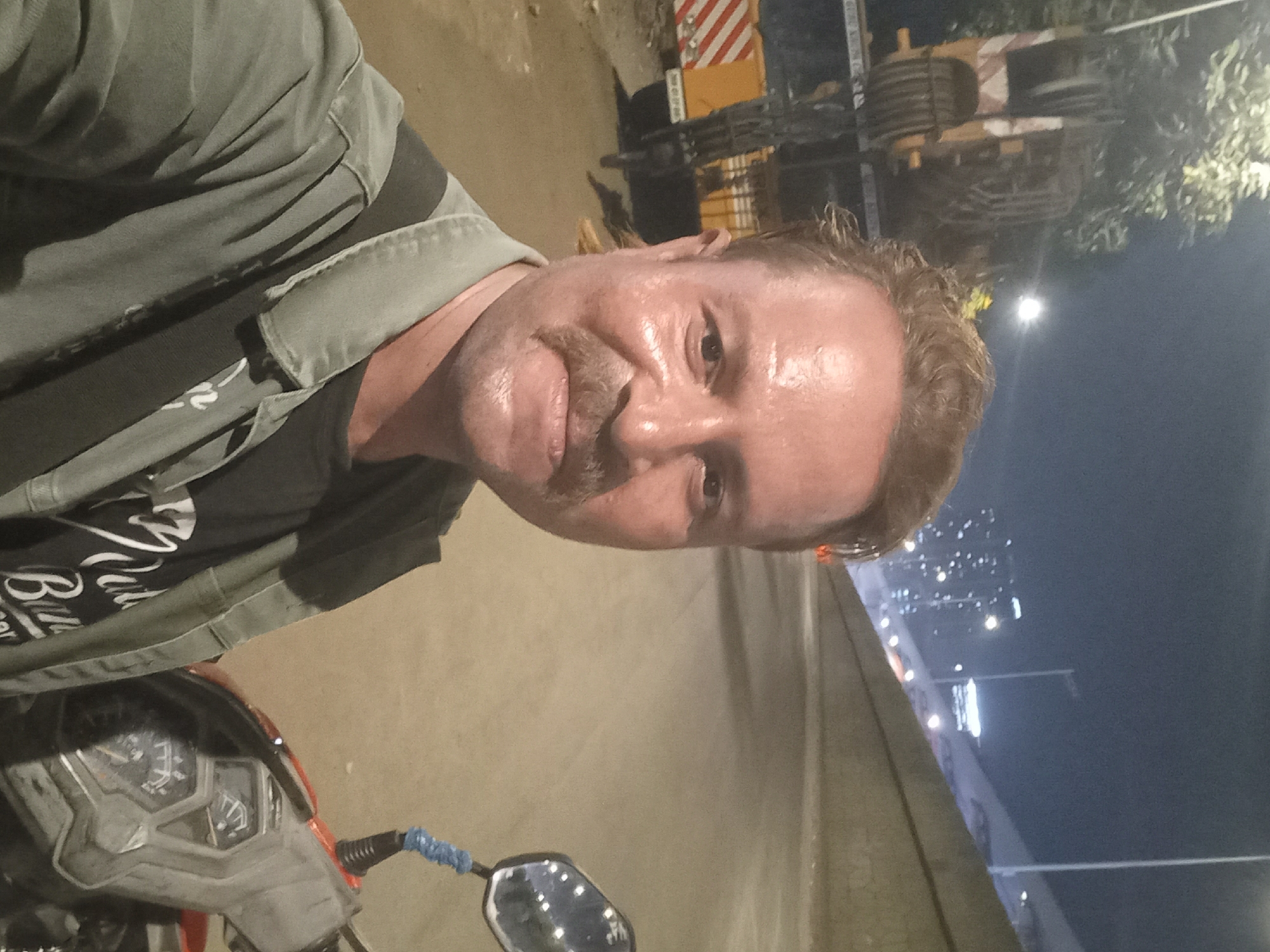 A man with light skin, reddish-brown hair, and a mustache is captured in a close-up selfie, slightly smiling. He appears to be sweating or his face is shiny. He is wearing a dark olive-green shirt over a black t-shirt. On the black t-shirt, partial white cursive text is visible, including "Bill..." and other letters.
The man is positioned on what appears to be a scooter or motorcycle, with parts of its dashboard, handlebars, and a rearview mirror visible in the lower left of the frame. A blue elastic band is wrapped around the handlebar grip.
The scene is set at night in an urban environment, likely in Mumbai, India, given the context. In the background, heavy machinery, possibly a construction crane with red and white striped warning markings, is visible under artificial lights. Distant city lights from buildings and streetlights illuminate the dark sky, suggesting a bustling area, possibly a road or flyover. The overall impression is a night-time snapshot of the man on his vehicle amidst city construction.