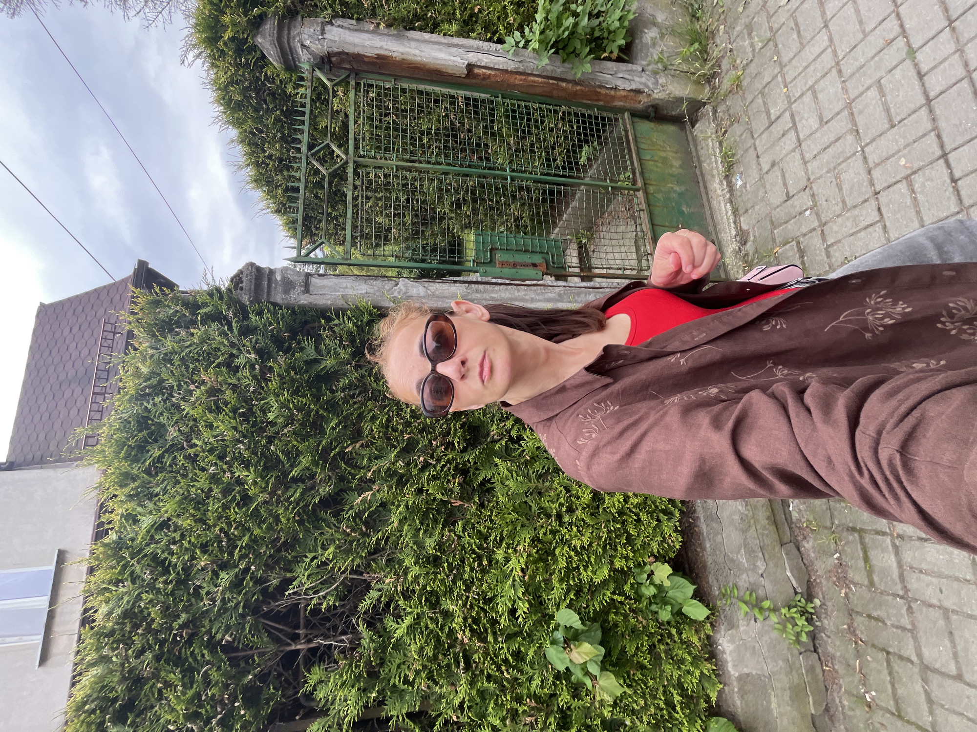 In an outdoor, eye-level medium shot, a woman with blonde hair pulled back and wearing sunglasses, a brown patterned shirt, and a red top stands on a paved walkway. She is positioned in the foreground, looking to her left and slightly away from the camera. Behind her, a tall, dense green hedge forms a backdrop, with a weathered, ornate green metal gate to her right. The gate is set between two rough-hewn stone posts. Beyond the gate and hedge, a sliver of a building with a dark, shingled roof is visible. The sky above is overcast.