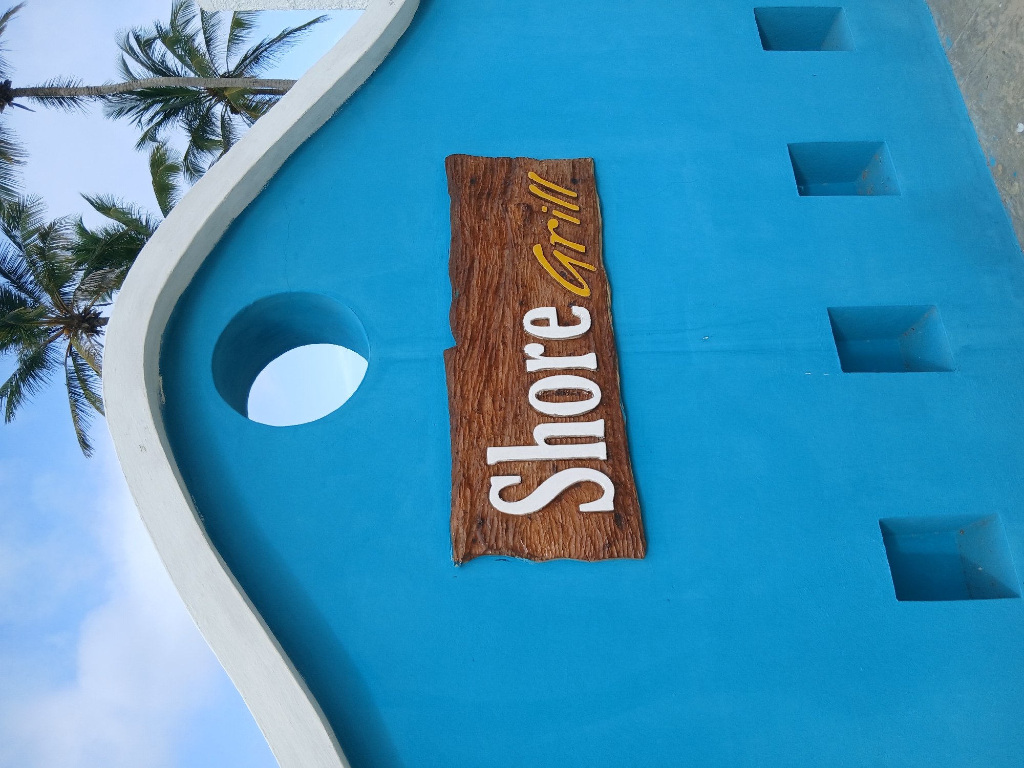 The image shows a close-up of a bright blue wall with a wooden sign that reads "Shore Grill". The sign has white lettering for "Shore" and yellow lettering for "Grill". Above the sign, there is a circular opening in the wall, and the wall has a white, curved trim along the top. Palm trees are visible against a blue sky with some clouds in the background. The lower part of the wall features three small, square recessed openings. The scene is set in Playa El Agua, Venezuela, suggesting a beachside or tropical location.