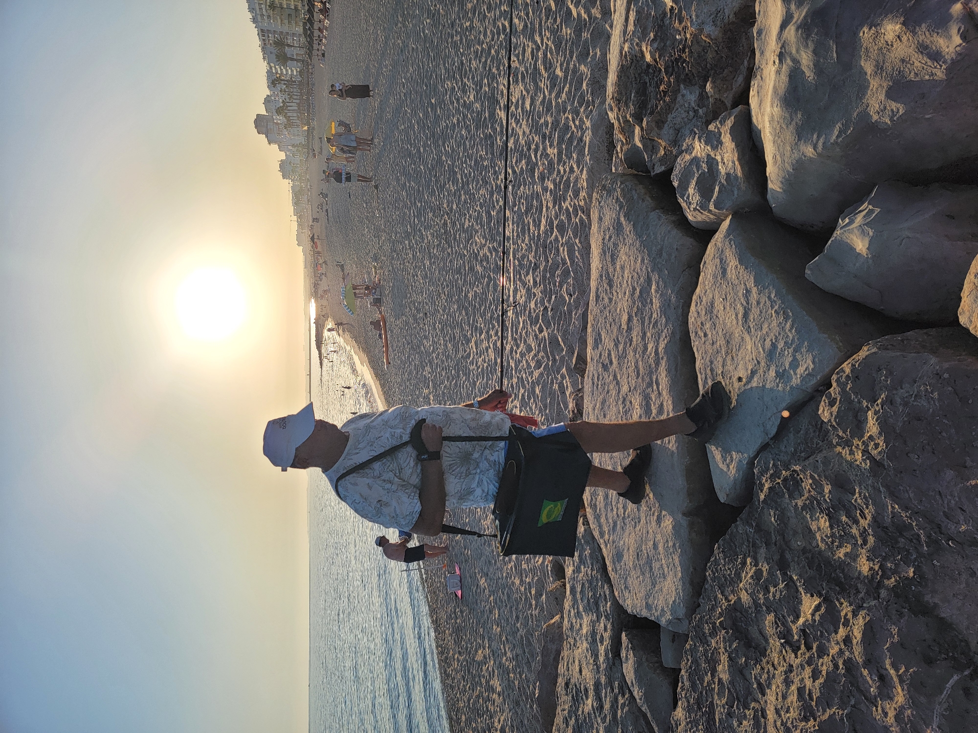 A man in a white cap, patterned shirt, and shorts walks on large rocks near the edge of a beach in Quarteira, Portugal. He carries a dark green bag with a strap over his shoulder and a fishing rod. The sun is low in the sky, casting a warm glow on the scene and illuminating the sandy beach, the ocean, and buildings in the distance. Other people are visible on the beach, some swimming in the water and others relaxing on the sand. The water is calm, and the sky is clear.