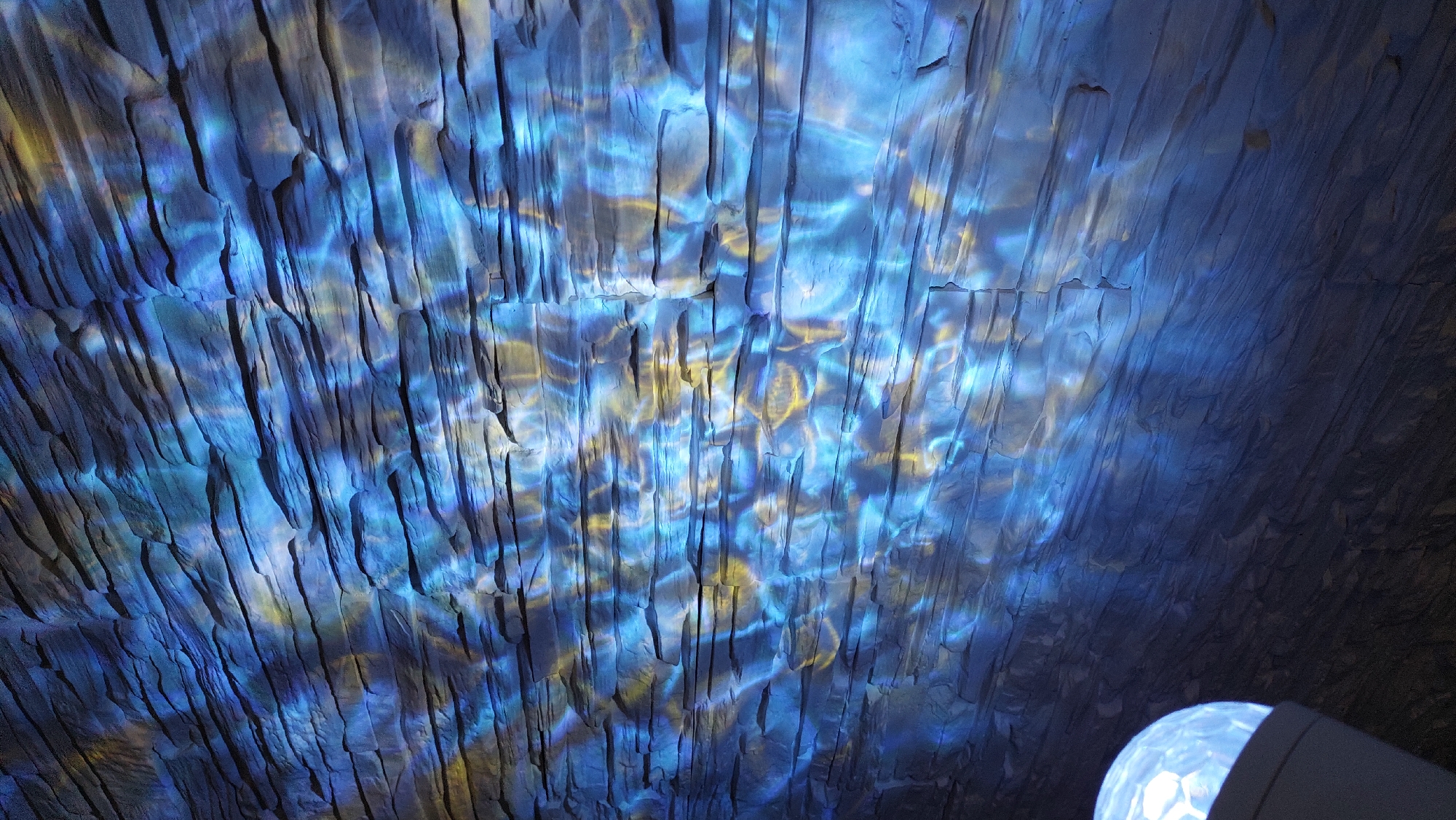 The image shows a ceiling with a rough, textured surface. The surface appears to be illuminated with blue and yellow light, creating a pattern of reflections and shadows. The texture of the ceiling is characterized by vertical lines or grooves. In the lower right corner of the image, a light source is visible. The location is Unknown, Unknown. There are no people or interactions visible in this image.