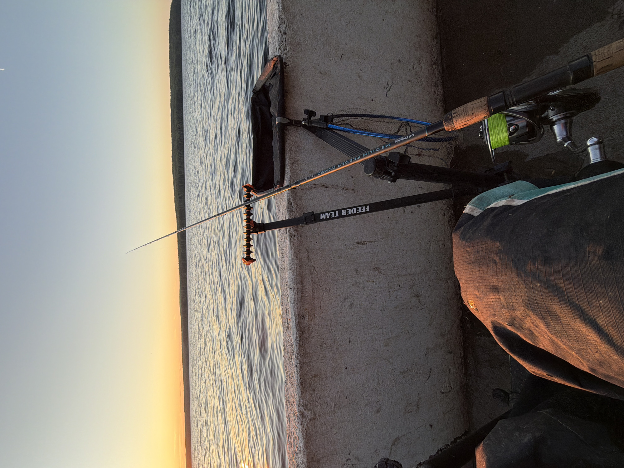 The image depicts a sunset scene at a body of water, likely the Kama River, in Chaikovsky, Russia. The foreground is occupied by a person's legs, clad in dark fishing pants, and various fishing equipment. Two fishing rods are propped up on stands, one of which has "FEEDER TEAM" written on it. A fishing net and a bait tray are also visible. The water is calm with gentle ripples, reflecting the warm colors of the sunset. The horizon is lined with a dense forest. The sky transitions from a pale blue near the top to a soft orange hue near the horizon. A sliver of a crescent moon is faintly visible in the upper right corner of the sky. The overall atmosphere suggests a peaceful evening spent fishing.