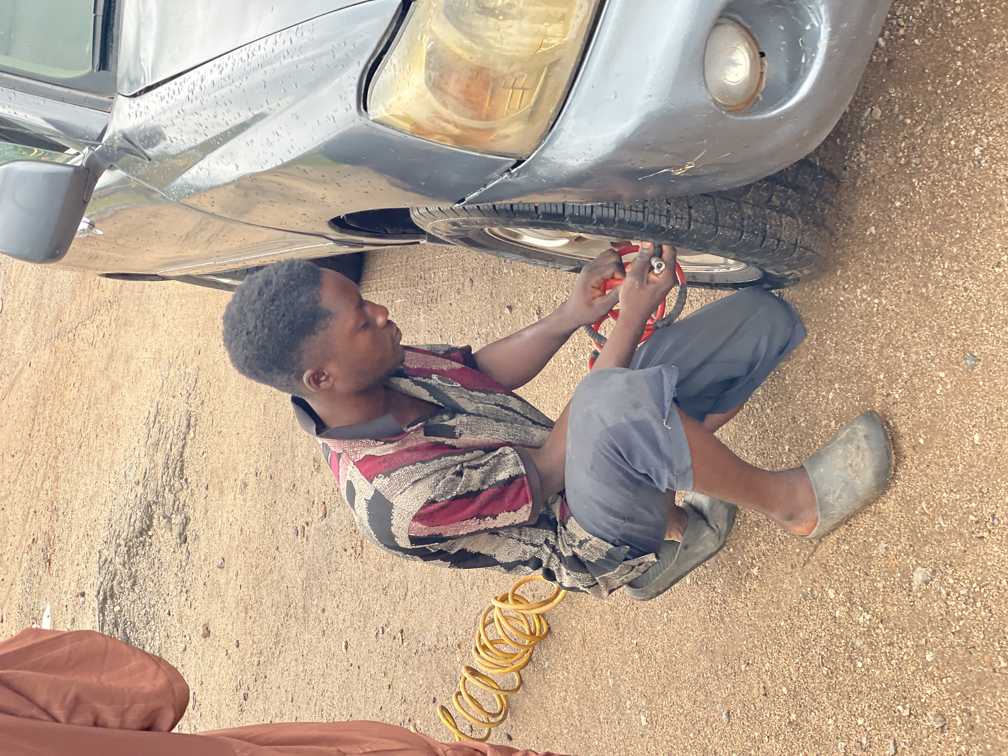 A man is shown working on the tire of a dark gray car. He is kneeling on the ground next to the front passenger-side tire, holding a red-handled tool connected to a coiled yellow hose. The man is wearing a patterned shirt with red, black, and white stripes, dark blue pants, and gray rubber shoes. The ground is covered in sand and small pebbles, suggesting an outdoor setting. The car's fender and headlight are visible, along with a reflection of the sky on the wet car body, indicating it might be raining or has recently rained. The scene appears to be in Lokoja, Nigeria. The time of day is not definitively indicated, but the lighting suggests it is daytime. There are no visible emotions or text in the image.