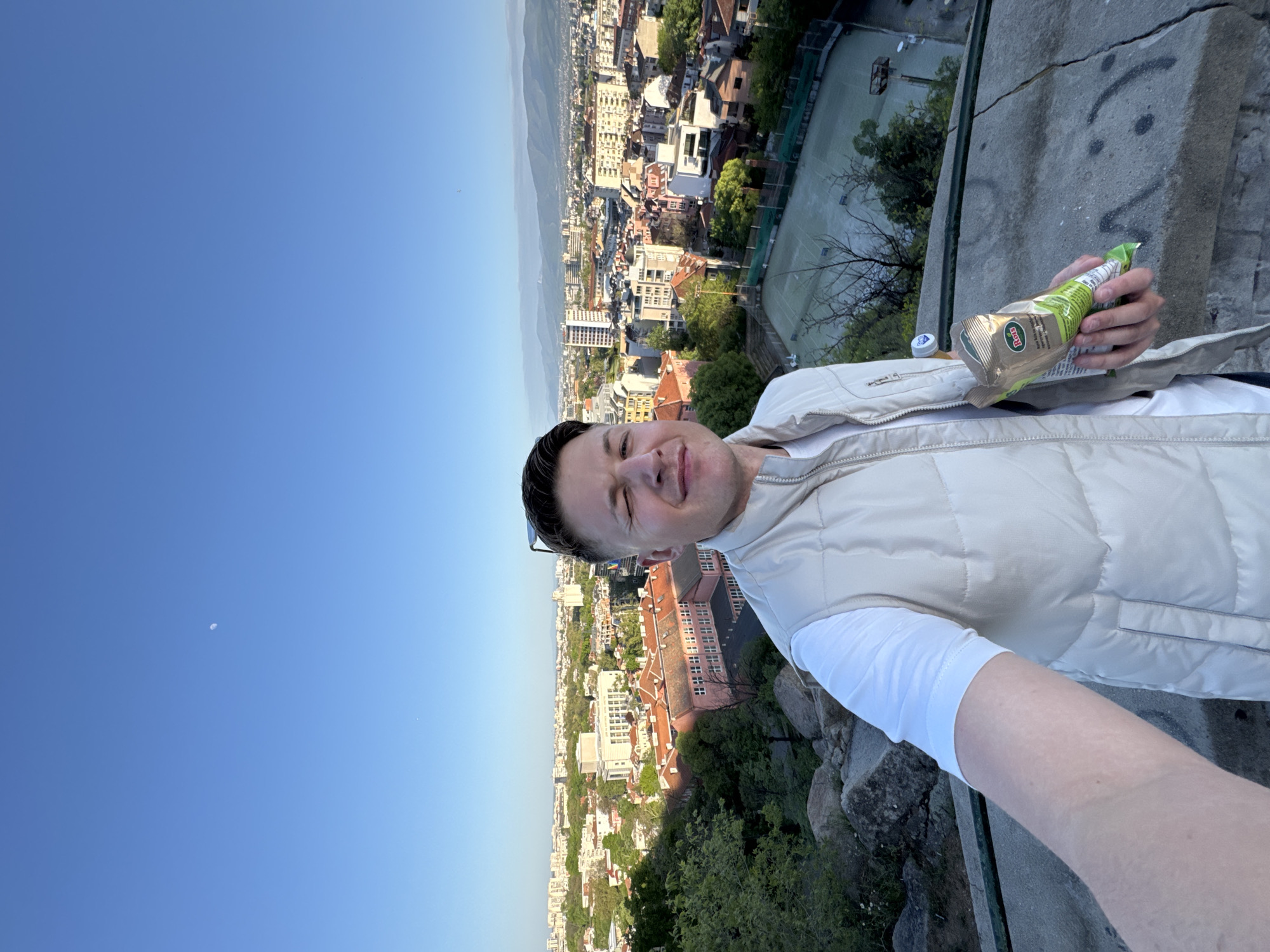 The image captures a man taking a selfie from a high vantage point overlooking the city of Plovdiv, Bulgaria. The photo is oriented 90 degrees counter-clockwise, showing the man and the cityscape on their sides.
The man, positioned in the foreground, smiles directly at the camera, conveying a sense of happiness or contentment. He wears a light beige or off-white puffer vest over a white long-sleeved shirt, with a pair of sunglasses pushed up onto his head. In his right hand, he holds a green and gold snack bag, partially crushed. Visible text on the bag includes the beginning of a brand name, possibly "Rö..." or "Ro...", and Bulgarian text "НАТУРАЛНИ..." (Natural...). A white plastic bottle cap rests on the concrete barrier next to his hand.
Behind the man, the sprawling cityscape of Plovdiv extends to the horizon under a clear, bright blue sky, indicating daytime, likely late afternoon or early evening. The city features a mix of buildings with various architectural styles, including many with reddish-brown roofs, interspersed with numerous green trees. In the distant background, faint mountain ranges complete the panoramic view. To the man's right, a sports court with a green surface, possibly for tennis or basketball, is visible within the urban fabric.
In the immediate foreground, a weathered concrete barrier separates the viewer from the city below. This barrier has some light graffiti, including what appears to be a simple smiley face. In the upper left corner of the expansive blue sky, a tiny white speck, possibly the moon, is discernible.