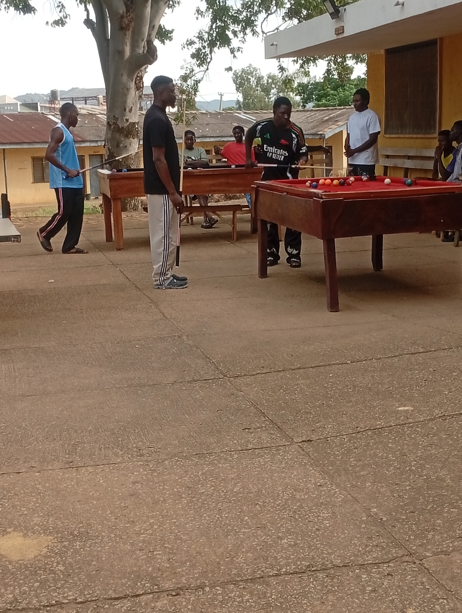 A group of young men are gathered around a pool table outdoors, possibly in a compound in Jos, Nigeria. The man in the foreground, wearing a black t-shirt and gray tracksuit pants, is standing near the pool table. To his left, another man in a blue tank top and black pants holds a pool cue. To the right of the man in the black t-shirt, a man in an "Emirates Fly Better" branded shirt is playing pool, aiming with a cue. Another man in a white shirt stands nearby, observing. Several colored billiard balls are scattered on the red felt of the pool table. The scene takes place on a paved outdoor area, with buildings and trees visible in the background. The lighting suggests it is daytime.