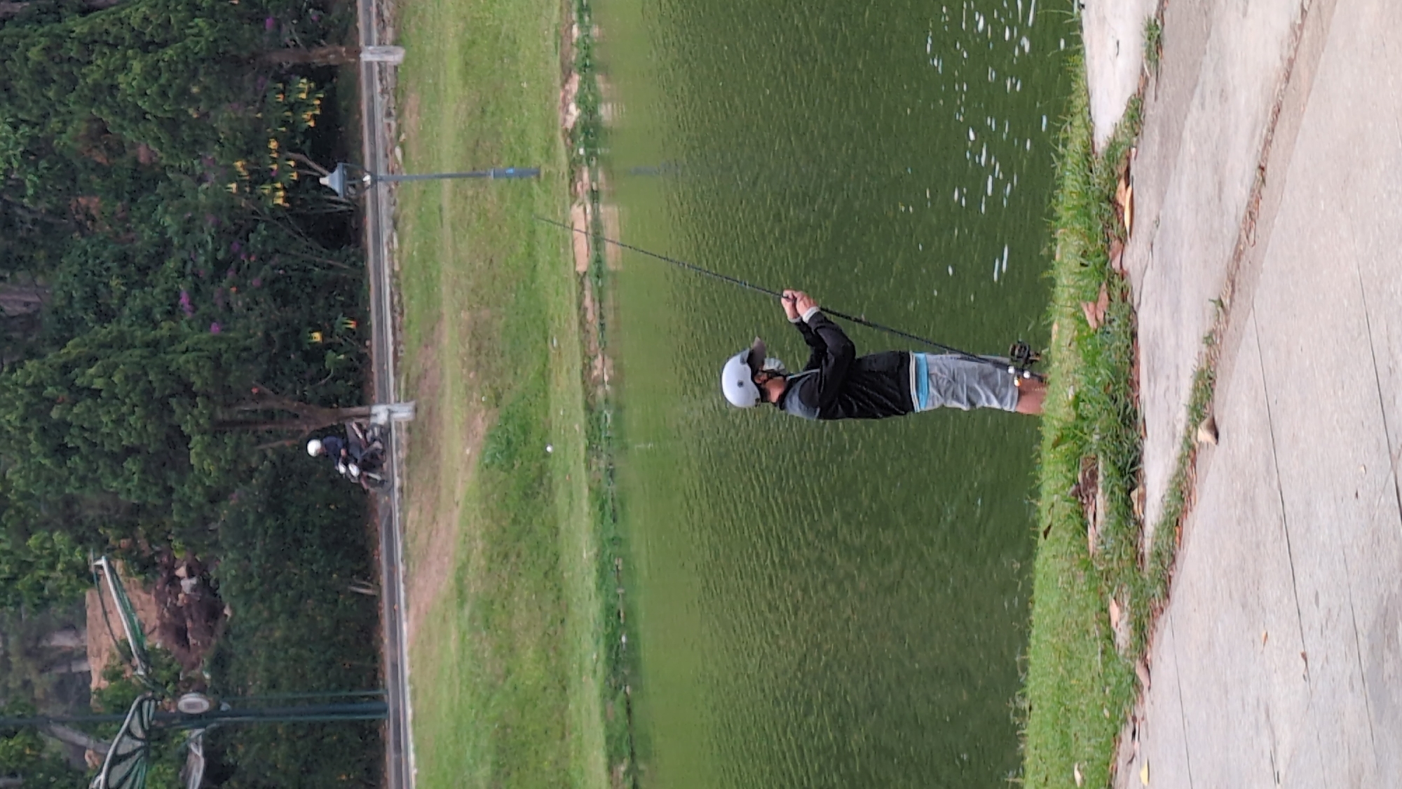 A man is fishing at Xuân Hương Lake in Đà Lạt, Vietnam. He stands on a paved walkway next to the lake, holding a fishing rod. He is wearing a black jacket, gray shorts, and a white helmet. The lake is green, and there are trees and grass on the opposite bank. A motorcycle is visible on the other side of the lake.