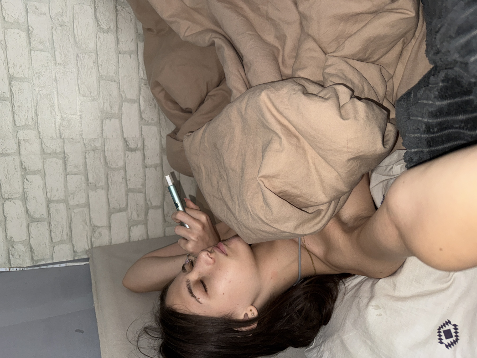 A young woman with dark hair is lying in bed, facing left, with her eyes closed. She is holding a light blue and white object in her right hand near her face. The woman is covered with a light brown blanket. The background shows a white brick wall. The scene appears to be in a bedroom.