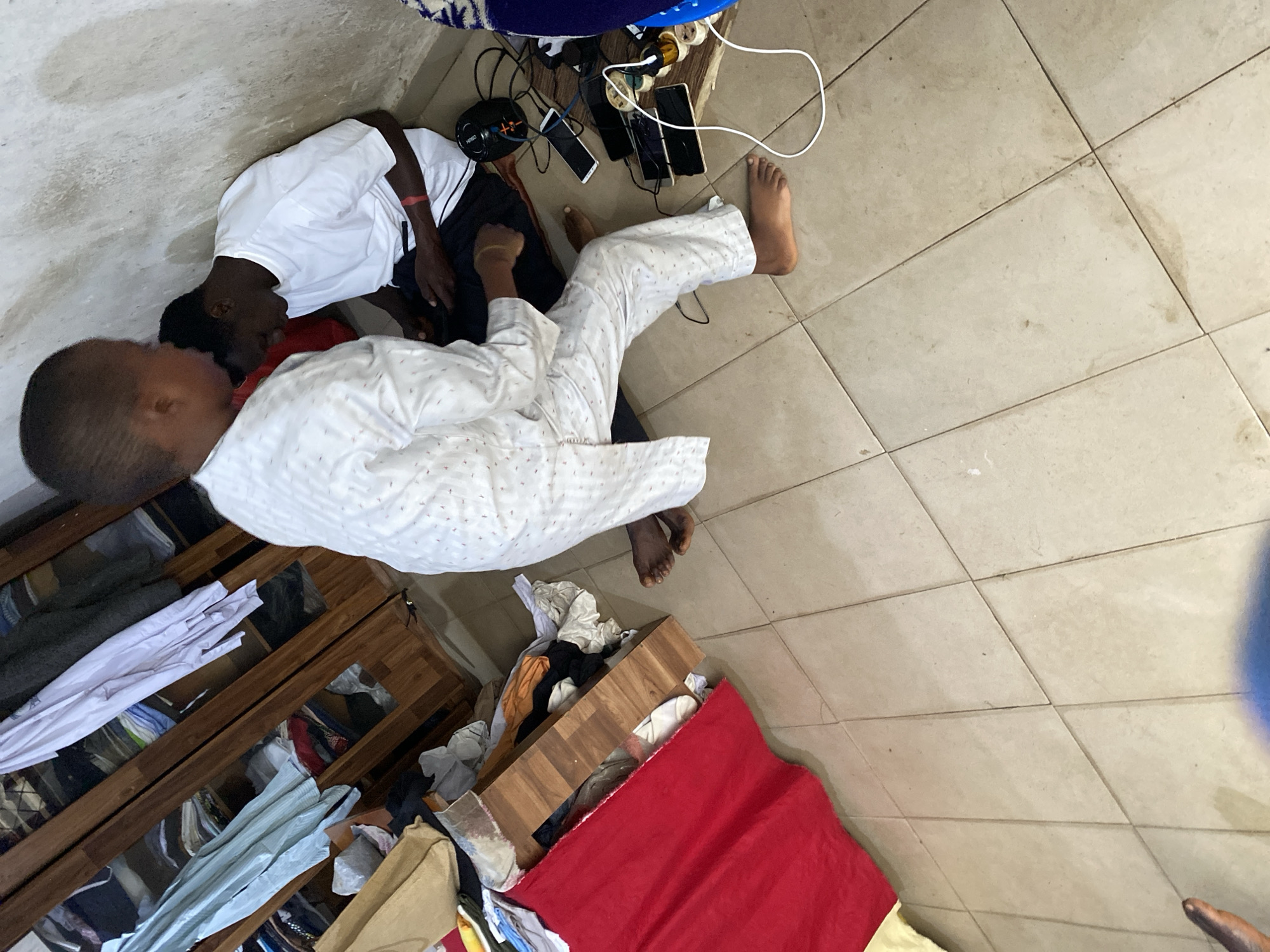 Two young males are seated on the floor in what appears to be a cluttered room. One male, facing away from the camera, wears a white t-shirt and dark shorts, and is looking down at a black device he is holding. He has a red band on his left wrist. The other male, facing towards the camera, is wearing a white patterned traditional garment and is seated with his legs extended. He is looking down at the device held by the first male. Numerous mobile phones are connected to a power strip via charging cables, suggesting they are charging. The environment shows a wooden wardrobe filled with clothing, a red cloth draped over a wooden surface, and tiled flooring. The scene is likely located in Jos, Nigeria, given the context of the request.