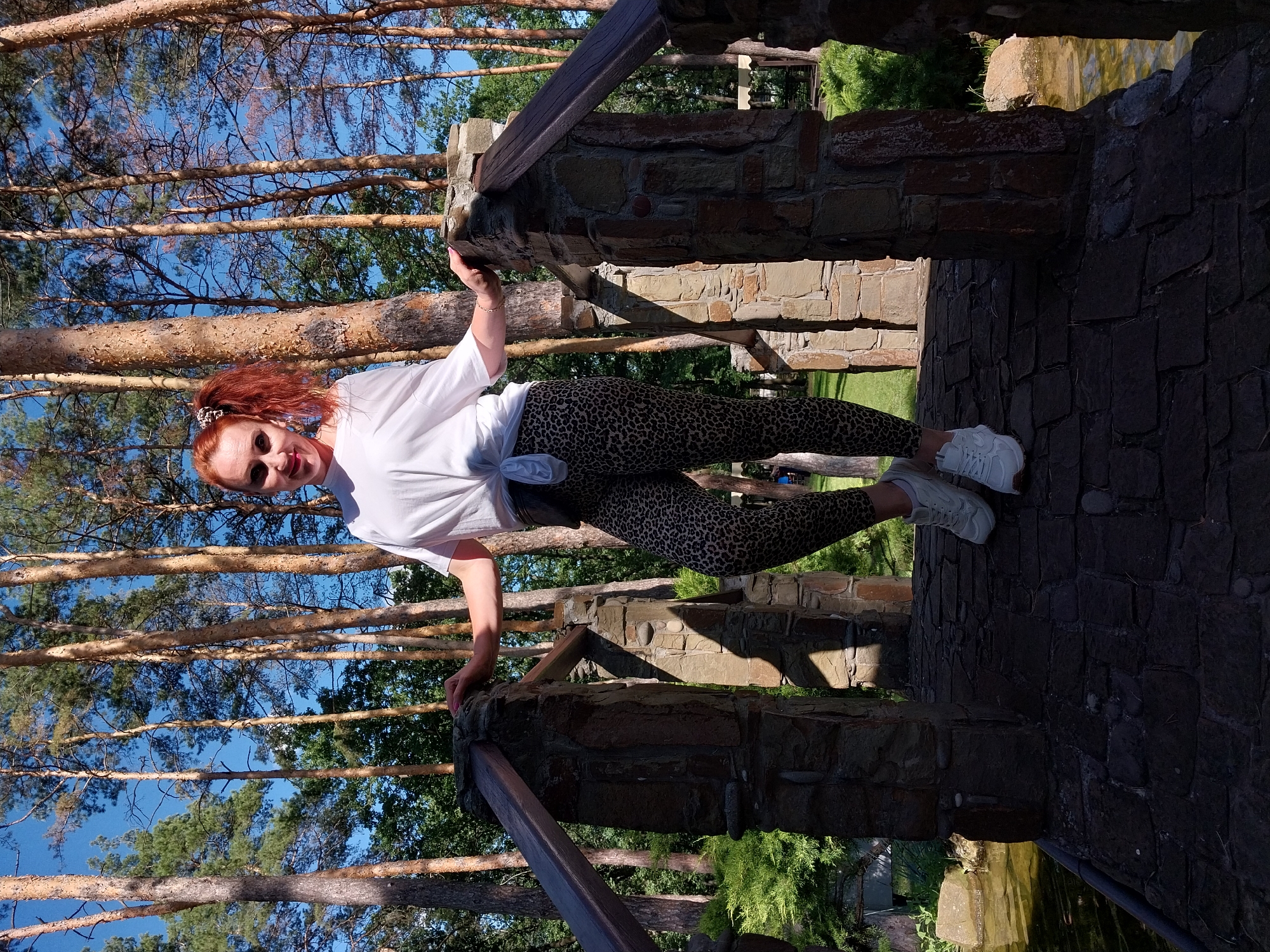 A woman with red hair pulled into a ponytail stands on a stone bridge in Orlivshchyna, Ukraine. She is wearing a white t-shirt tied at her waist, leopard-print leggings, and white sneakers. She is resting her hands on the stone railing of the bridge. The bridge is surrounded by trees and lush greenery. The sky is blue.
