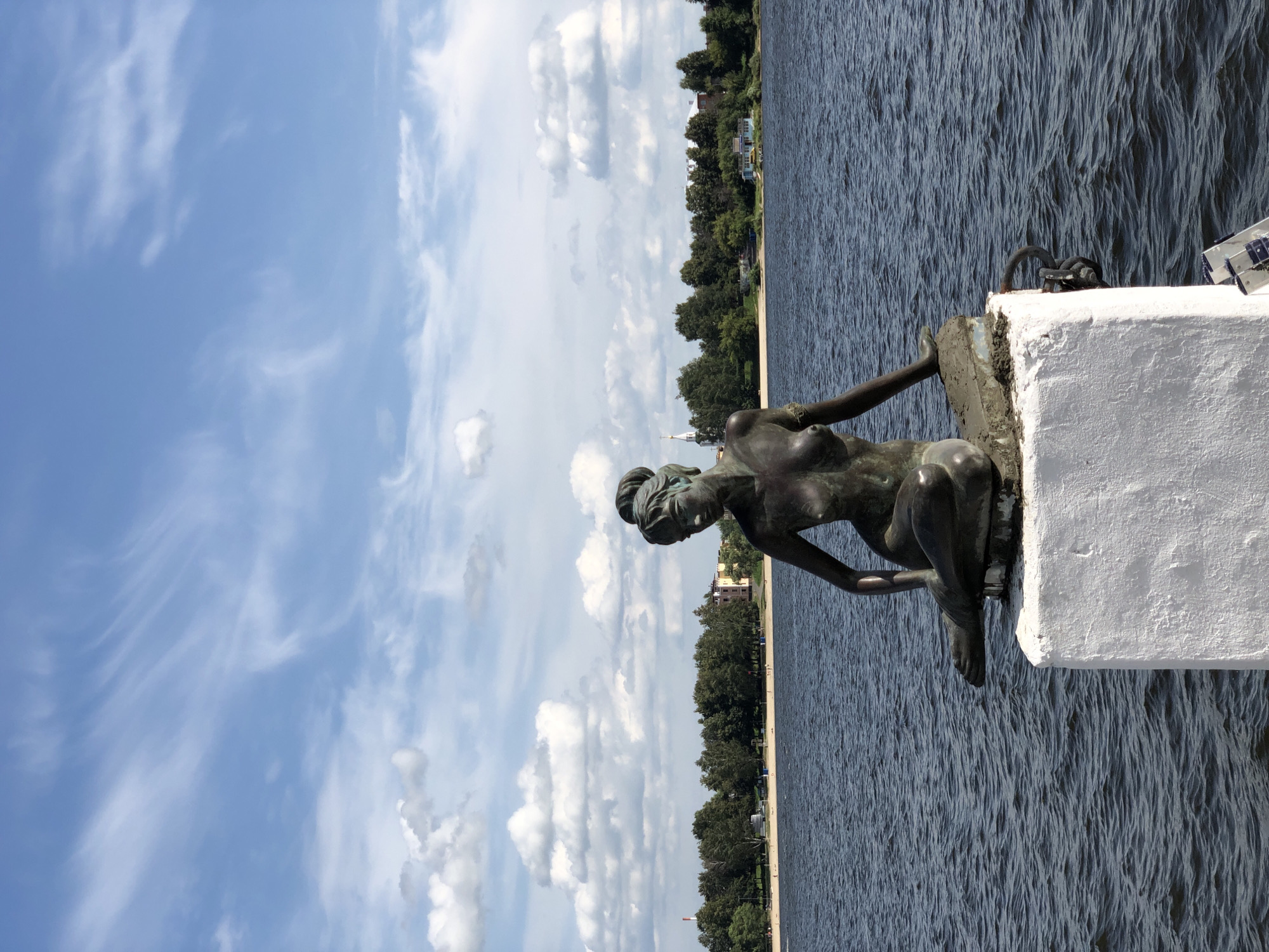 A bronze statue of a mermaid sits on a concrete pedestal in the water. The mermaid is depicted in a seated position, with her legs crossed. The statue is dark in color and appears to be made of aged bronze. The pedestal is white and rectangular, with some grime and wear visible. The water is dark blue and appears choppy, with small waves. In the background, there is a sandy beach lined with trees. Beyond the trees, buildings and a church spire can be seen. The sky is blue with scattered white clouds. This statue is located on the Volga River embankment in Yaroslavl, Russia, near the Strelka.