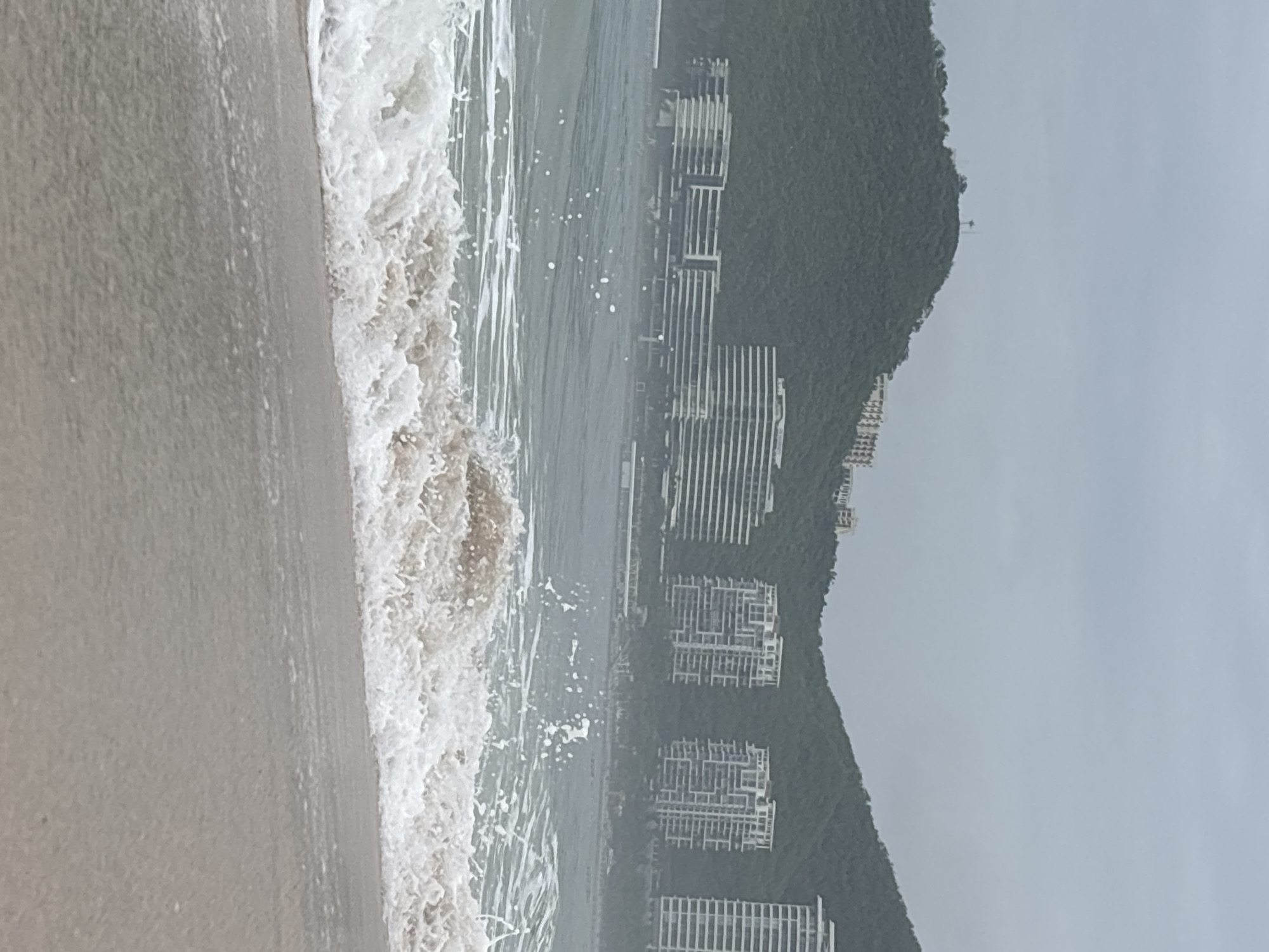 The image depicts a coastline scene with a body of water, a sandy beach, and hills in the background. The water appears to be a choppy sea with waves breaking near the shore, creating white foam. Several modern buildings with multiple stories and horizontal window patterns are visible on the hills in the distance. The sky is overcast, suggesting a cloudy or hazy day. The foreground of the image shows the wet sand of the beach where a wave is receding. There are no people or animals visible in the frame. The overall impression is of a coastal urban or resort area.